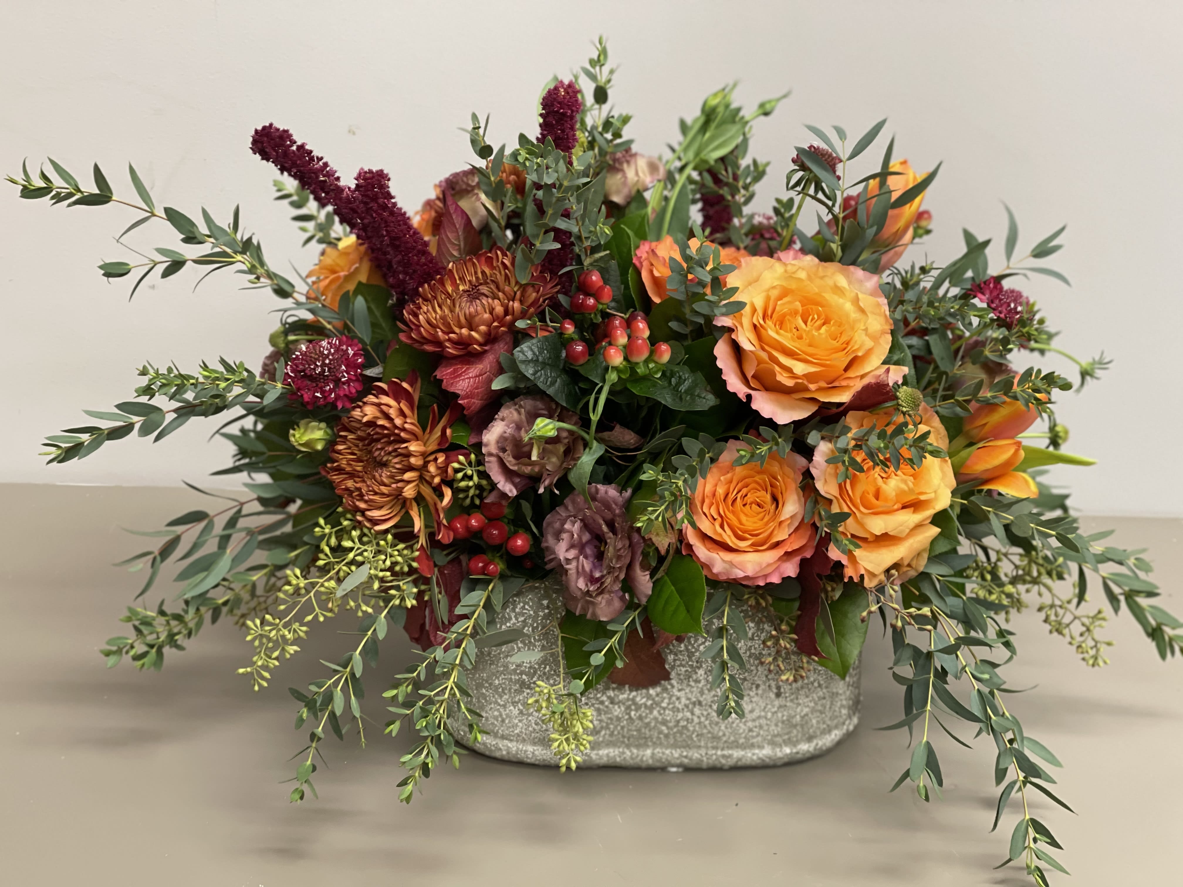 Low centerpiece of orange roses and rust mums with red accents in a gray ceramic container