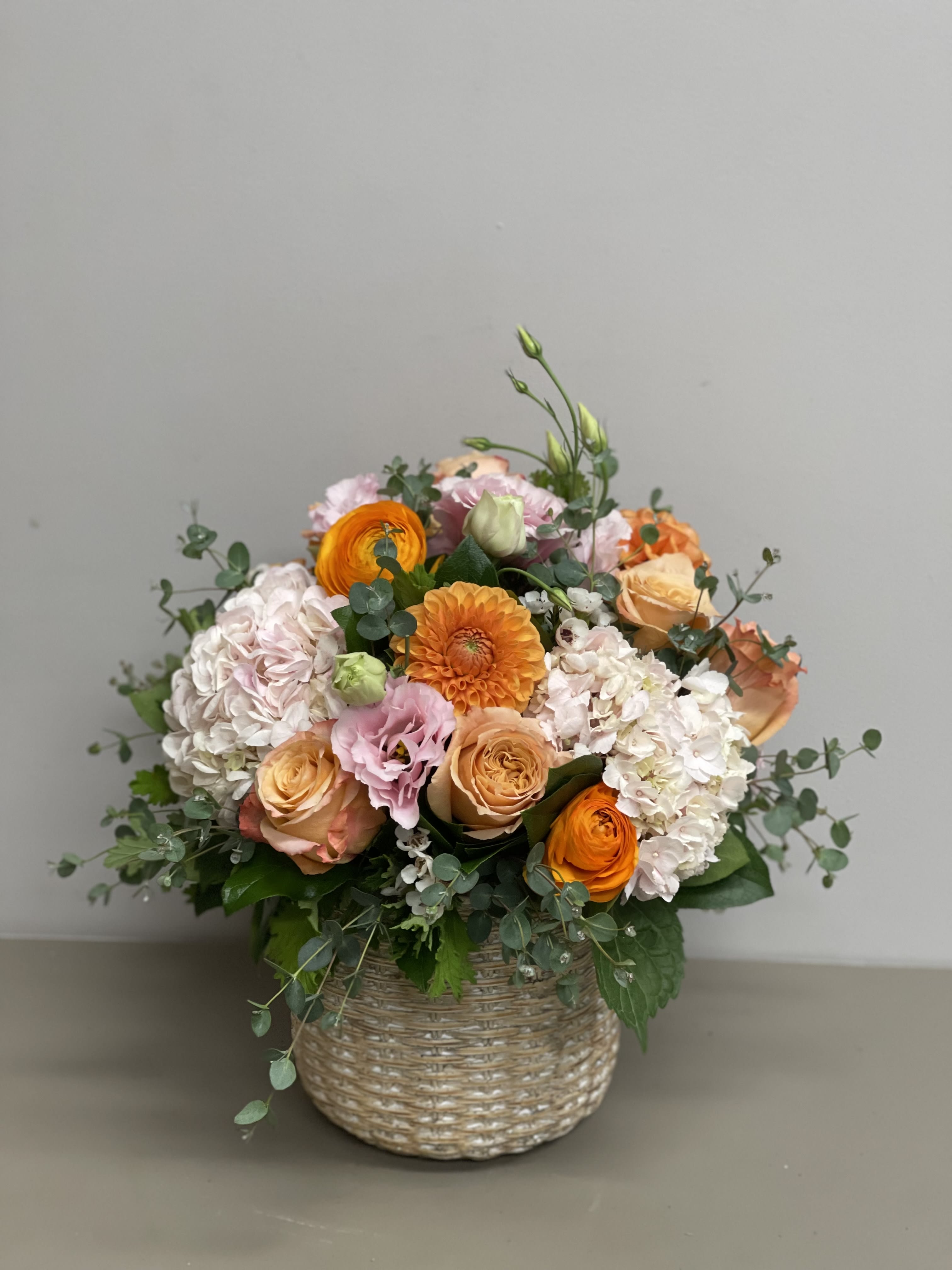 Round arrangement of peach and orange roses, hydrangeas, and pink blooms in a woven basket