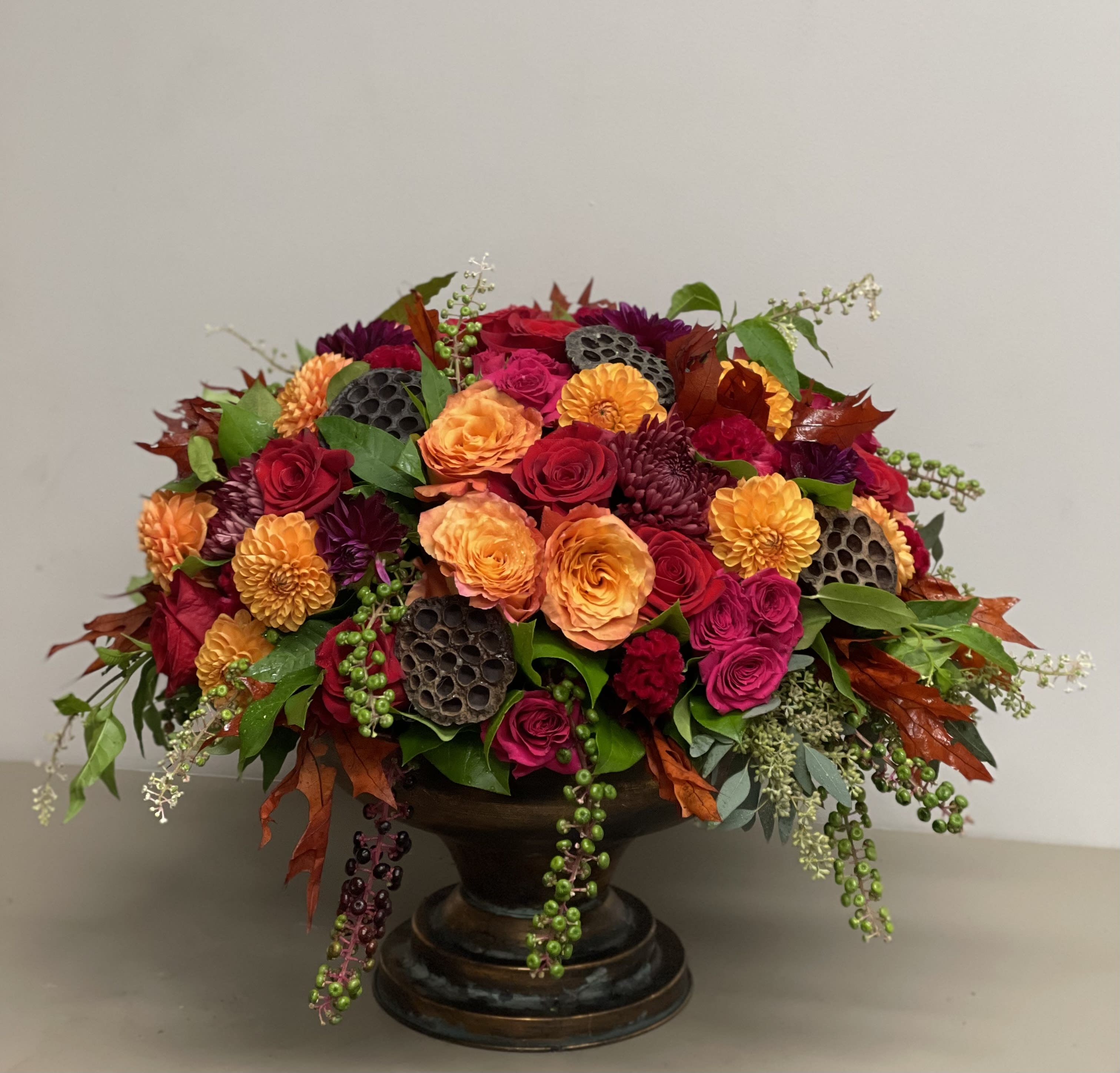Low arrangement of orange and red roses with dark accent flowers in a bronze pedestal bowl