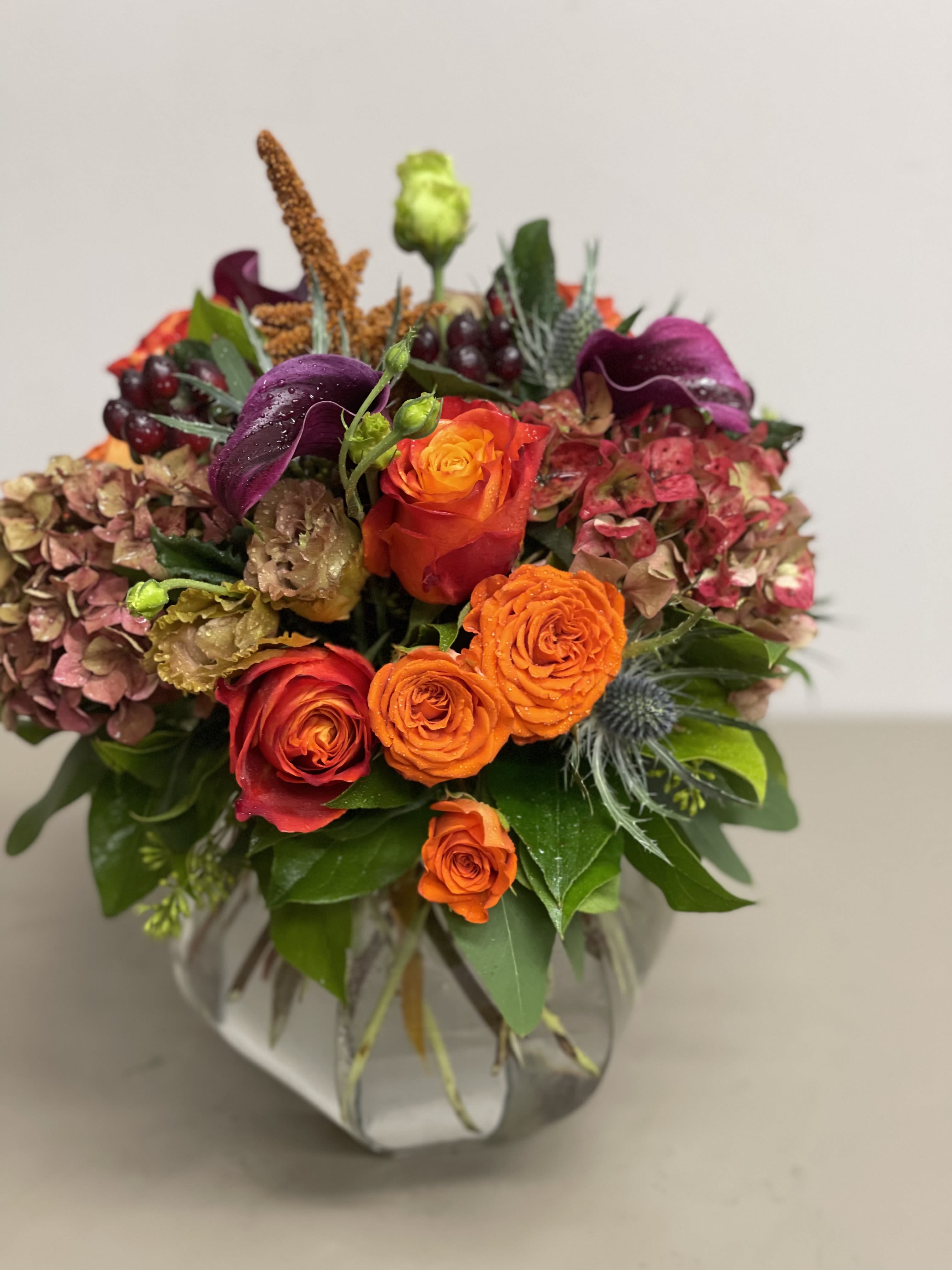 Low glass vase arrangement with bright orange and red roses, purple calla lilies, and antique hydrangeas