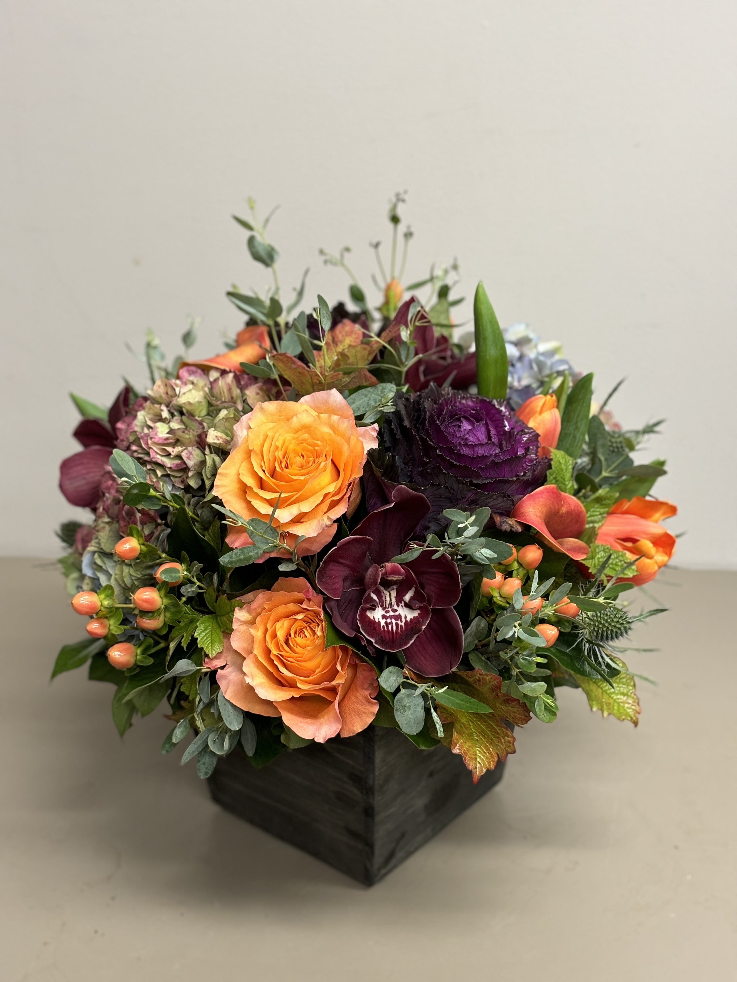 Low arrangement of orange roses, dark burgundy orchids, and mixed blooms in a square dark wooden vase