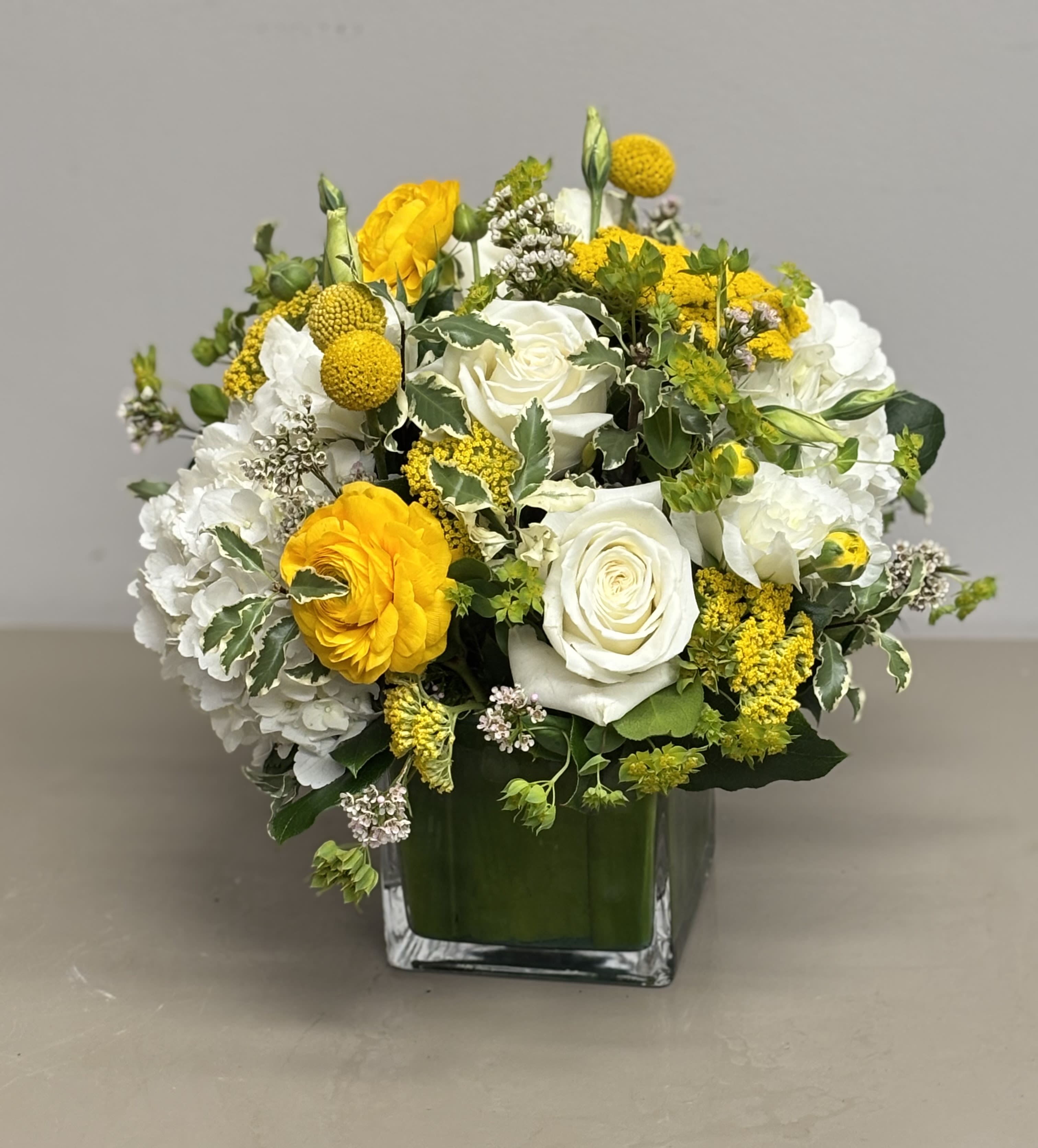 Rounded arrangement of white roses, hydrangeas, and yellow blooms in a clear square glass vase