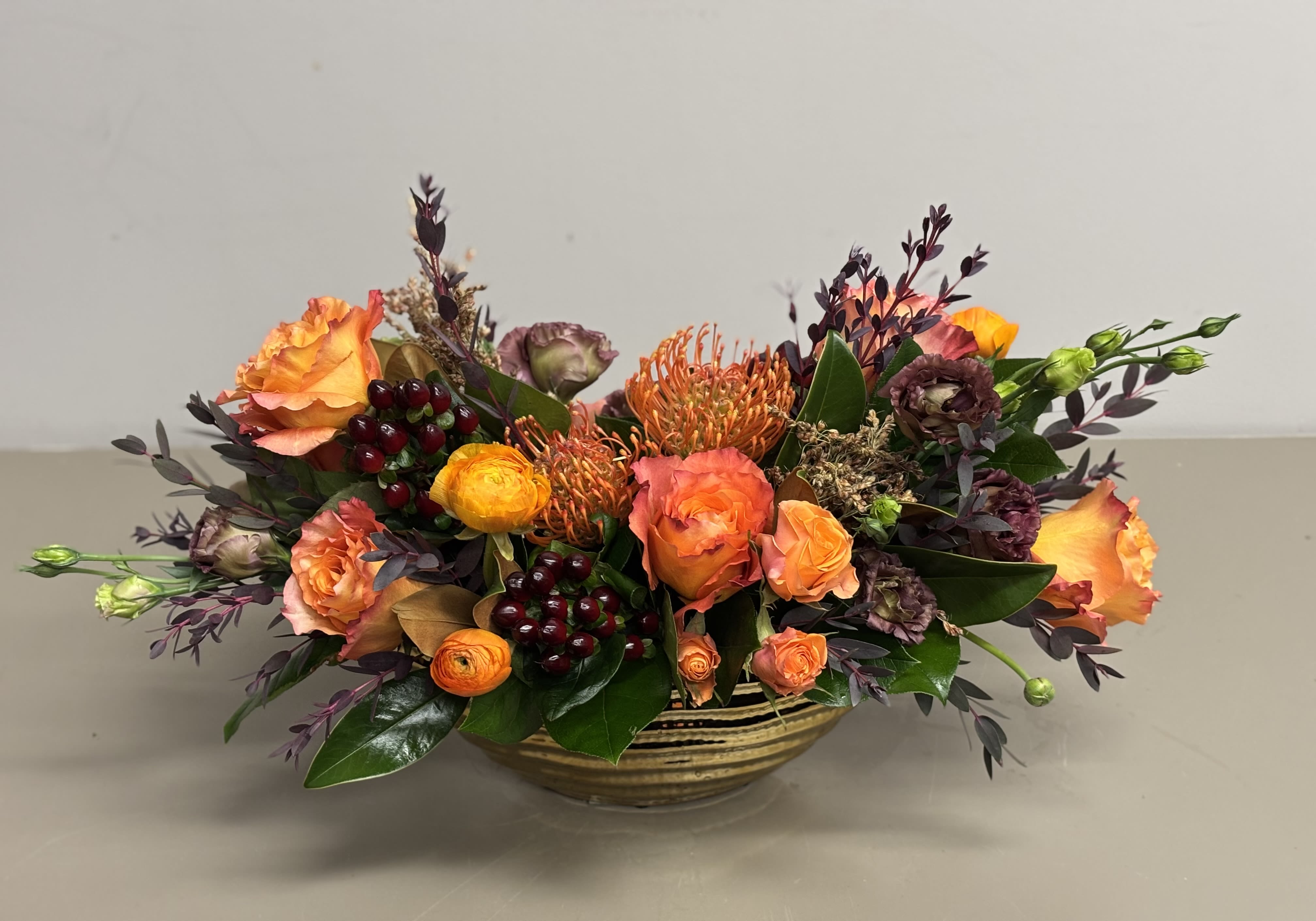 Low centerpiece of orange roses, pincushion protea, and dark berries in a striped gold bowl
