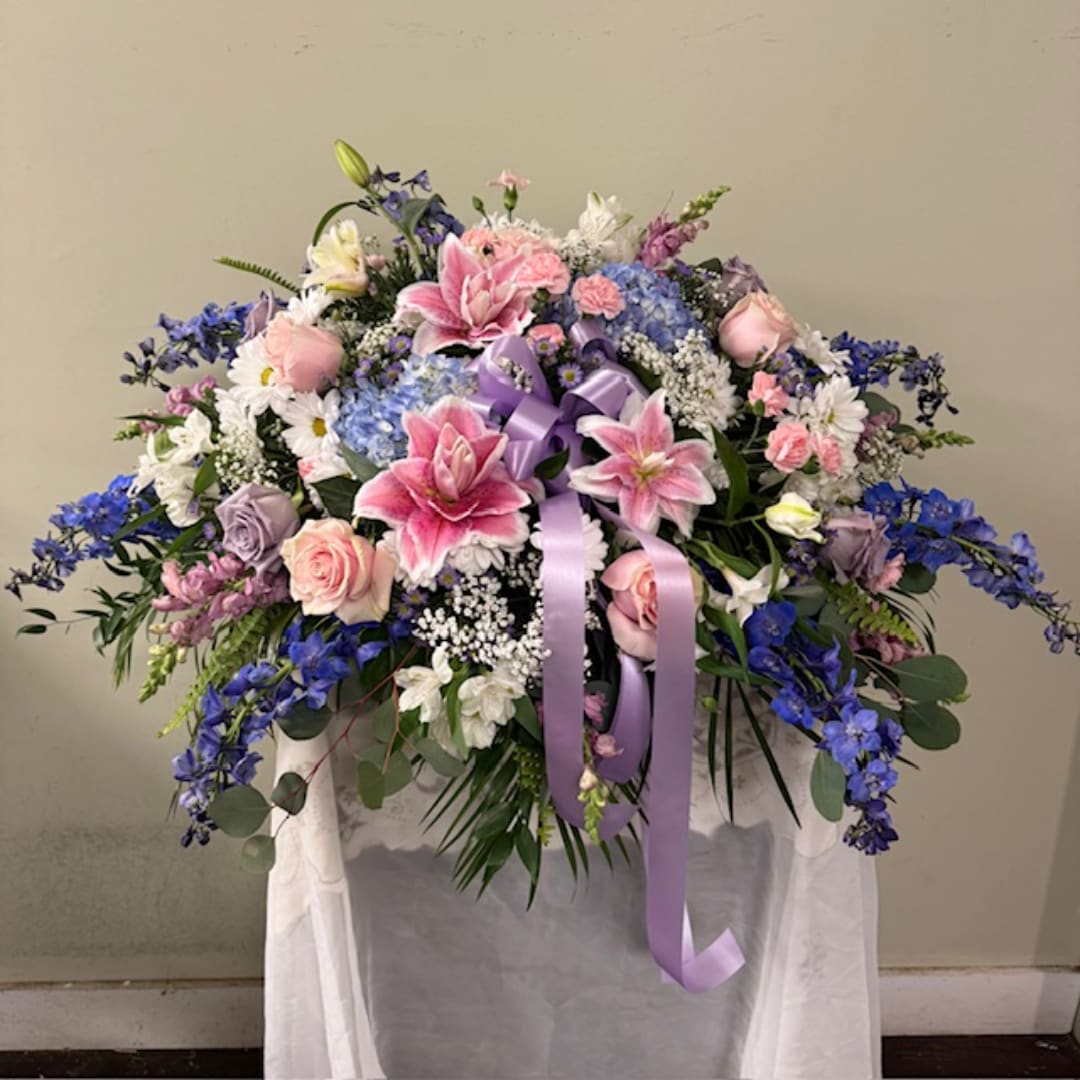 Large pastel floral arrangement with pink lilies, roses, and blue flowers on a draped stand