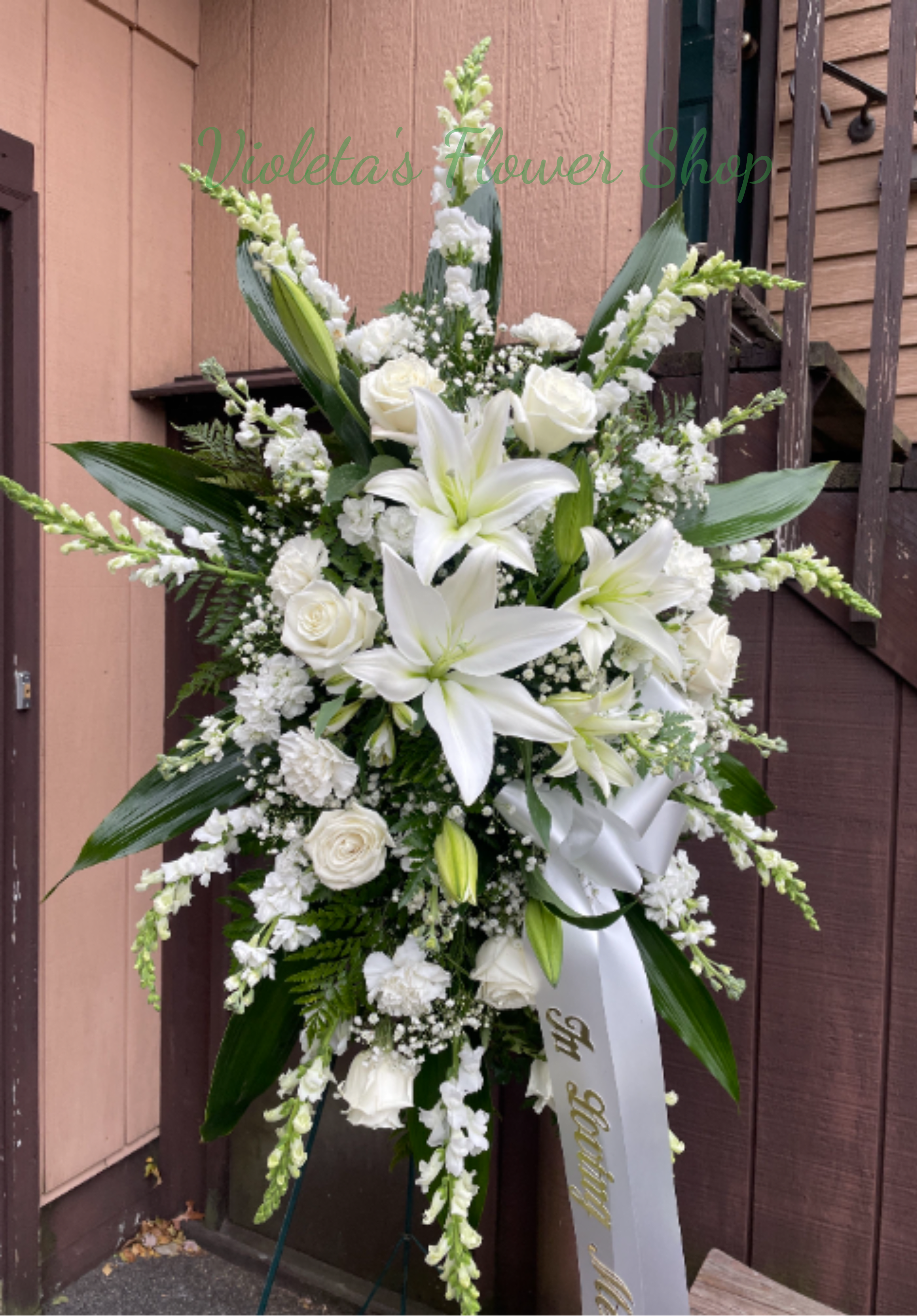 Memorable Tribute Standing Spray. - Towering white lilies and roses create a graceful, solemn statement in this large standing spray-perfect as a sympathy arrangement or funeral tribute. The tall, symmetrical design with long arching lines and broad dark green foliage frames the flowers so the person you're honoring will feel respected and remembered.  White carnations, vertical snapdragons and clouds of baby's breath add texture and gentle movement, while the clean white ribbon with gold lettering reinforces the formal memorial tone. This arrangement arrives ready to display on the included standing easel; its layered silhouette and mixed white blooms read clearly from a distance for chapel or graveside service. 
