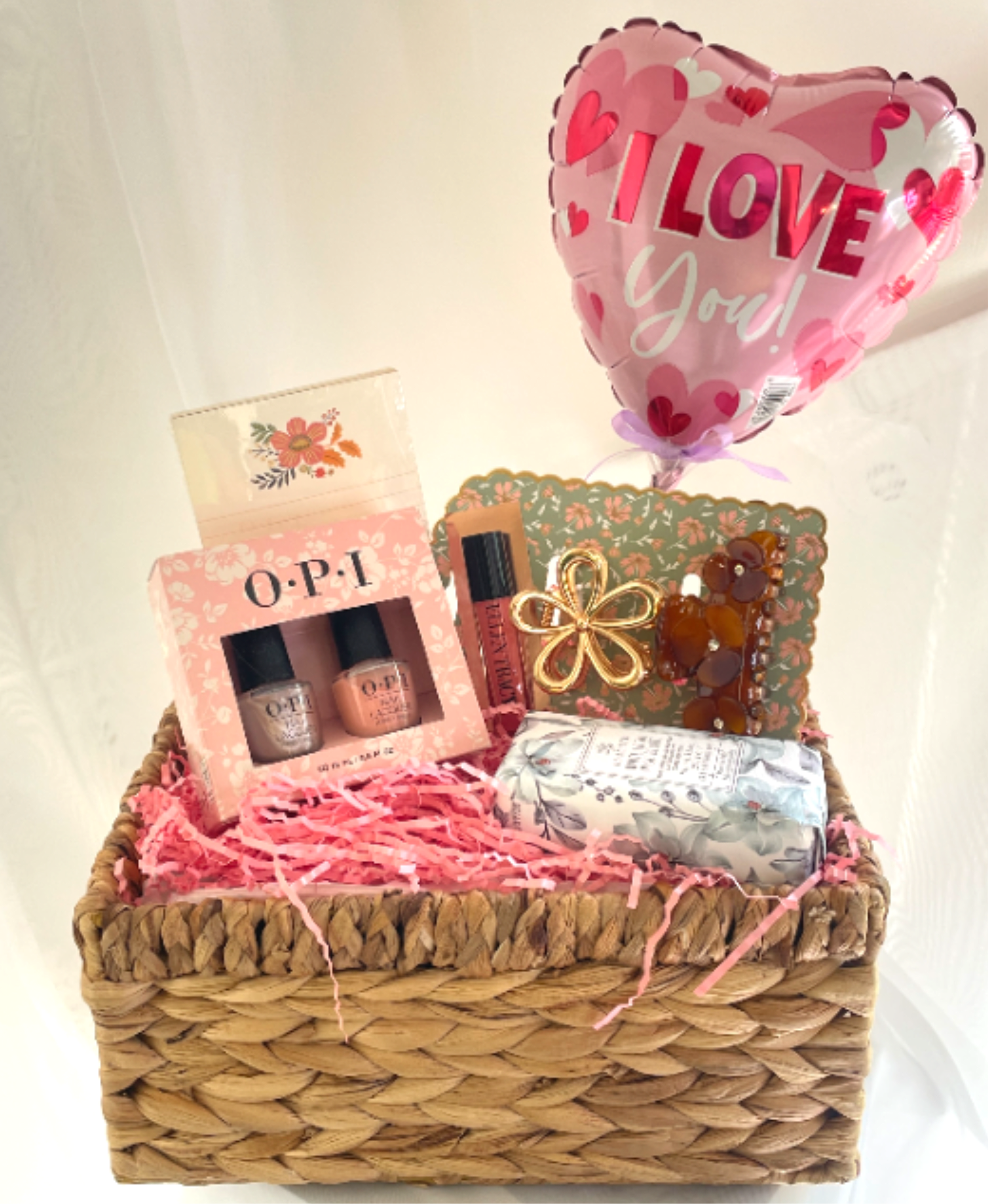 Gift Basket for Her - A cheerful woven gift basket arranged on a bed of pink shredded paper-this gift basket for her is a ready-made Thinking of You present that reads as warm, feminine, and slightly romantic. Topped with a pink heart-shaped foil balloon that says "I LOVE You!", it's perfect for a birthday, a sweet surprise, or a Valentine's note to the person you're thinking of who deserves a little pampering.  Inside you'll find a boxed OPI nail polish set, a lip gloss tube, a wrapped decorative soap bar, a gold flower-shaped claw hair clip, and a floral-patterned decorative card, all nestled in a rectangular woven basket with ribbon and a decorative box accent. The palette of pink, cream, and warm tan creates a soft, affectionate look; shredded paper filler keeps everything presented beautifully and secure.  