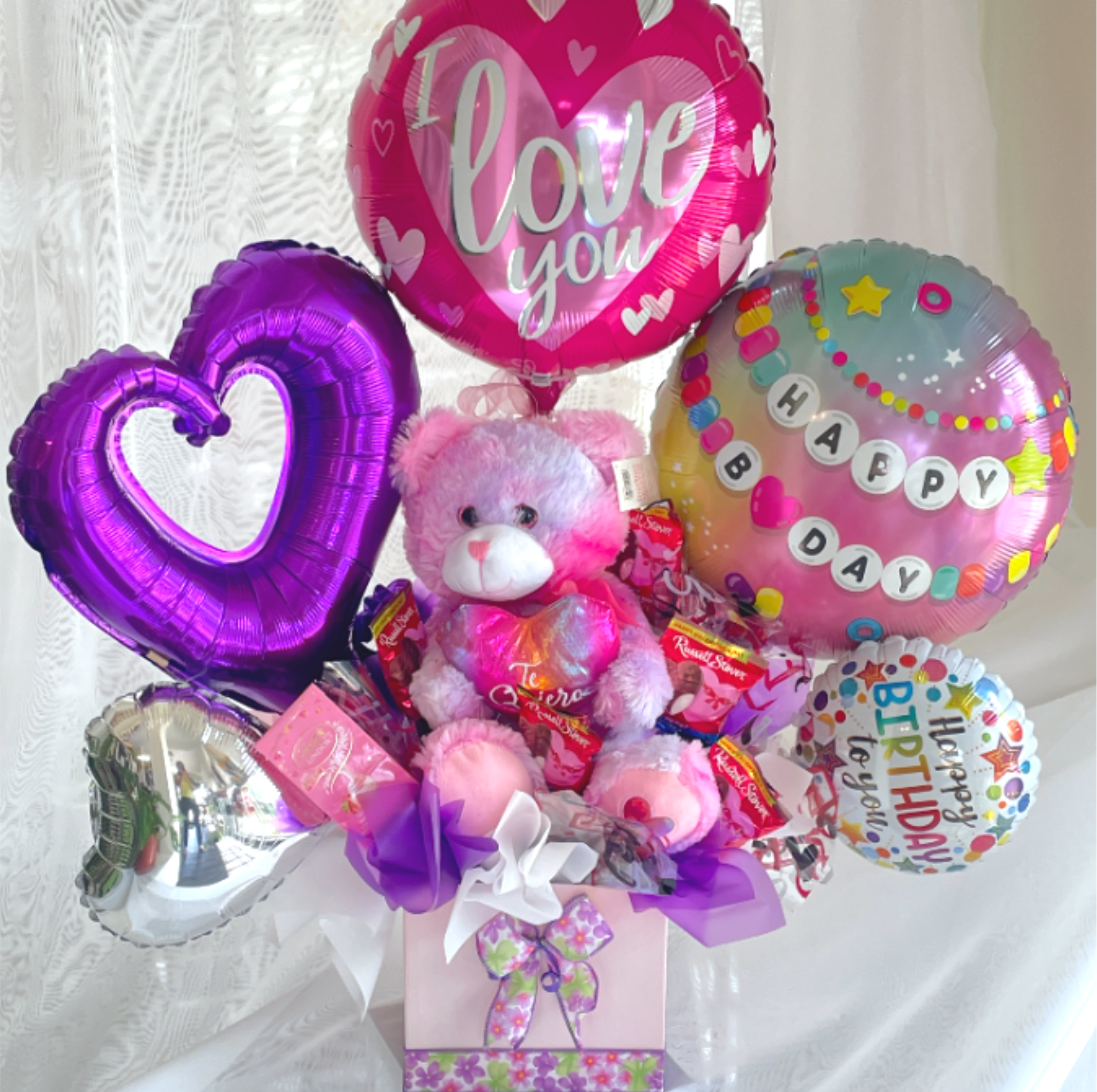 Birthday Gift Box - Bright pink and purple foil balloons, a cuddly pink teddy bear, and an assortment of chocolates and candy tucked into a pastel gift box-this birthday gift box makes the recipient feel celebrated and cherished from the moment they see it. Designed as a birthday or romantic surprise, the ensemble's "I love you," heart, and "Happy Birthday" balloons pair with ribbon bows for a playful, affectionate presentation that reads as both festive and intimate.  