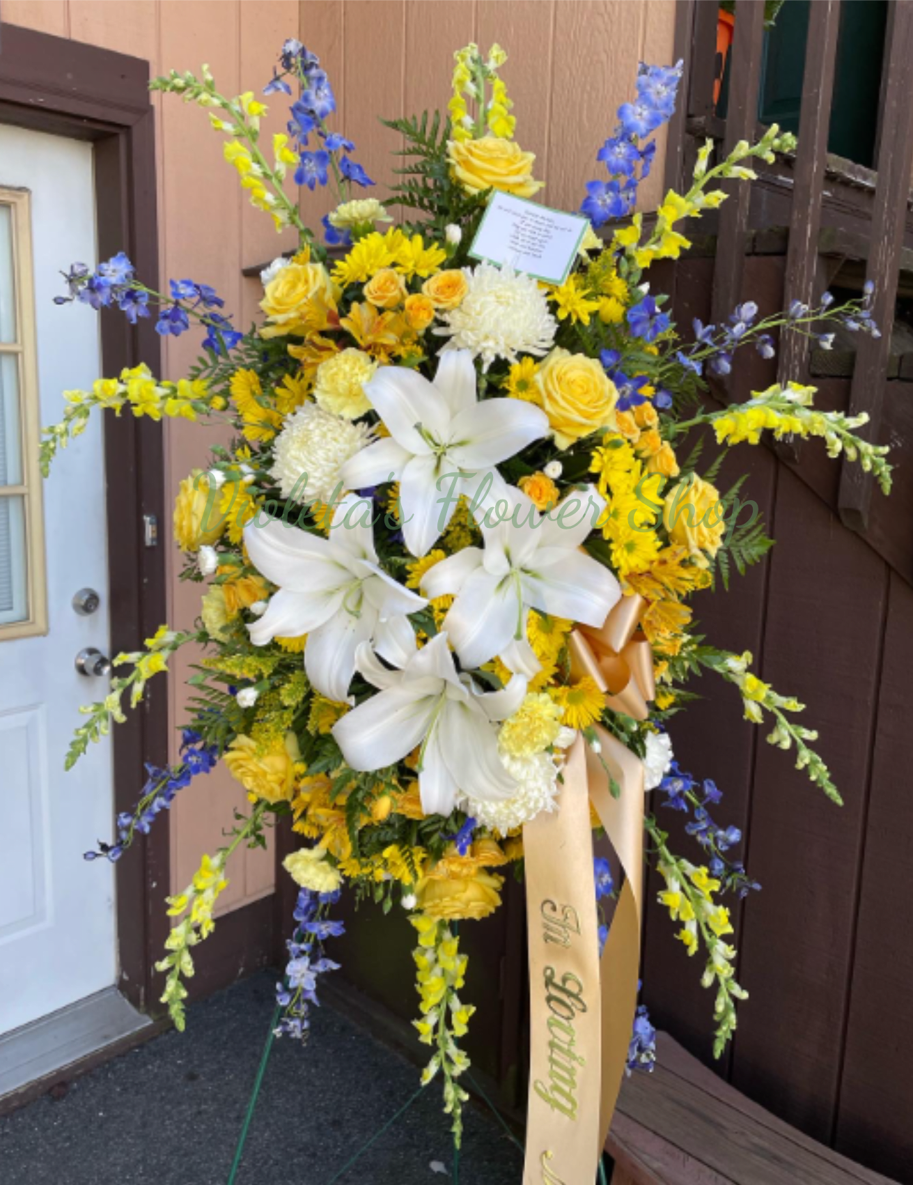 Golden Memories Standing Spray - This tall spray combines lilies, roses, chrysanthemums, snapdragons, daisies, alstroemeria and seasonal filler in a full, symmetrical composition on a standing easel. Long arching stems give the piece dramatic height while a pale ribbon and attached card complete the presentation. The palette of yellow, white, blue and green balances warmth and calm so the person you're honoring will feel remembered and celebrated.  Sized for visibility at services and graveside, this arrangement arrives ready on a standing easel for immediate display; include your message on the card when ordering. 