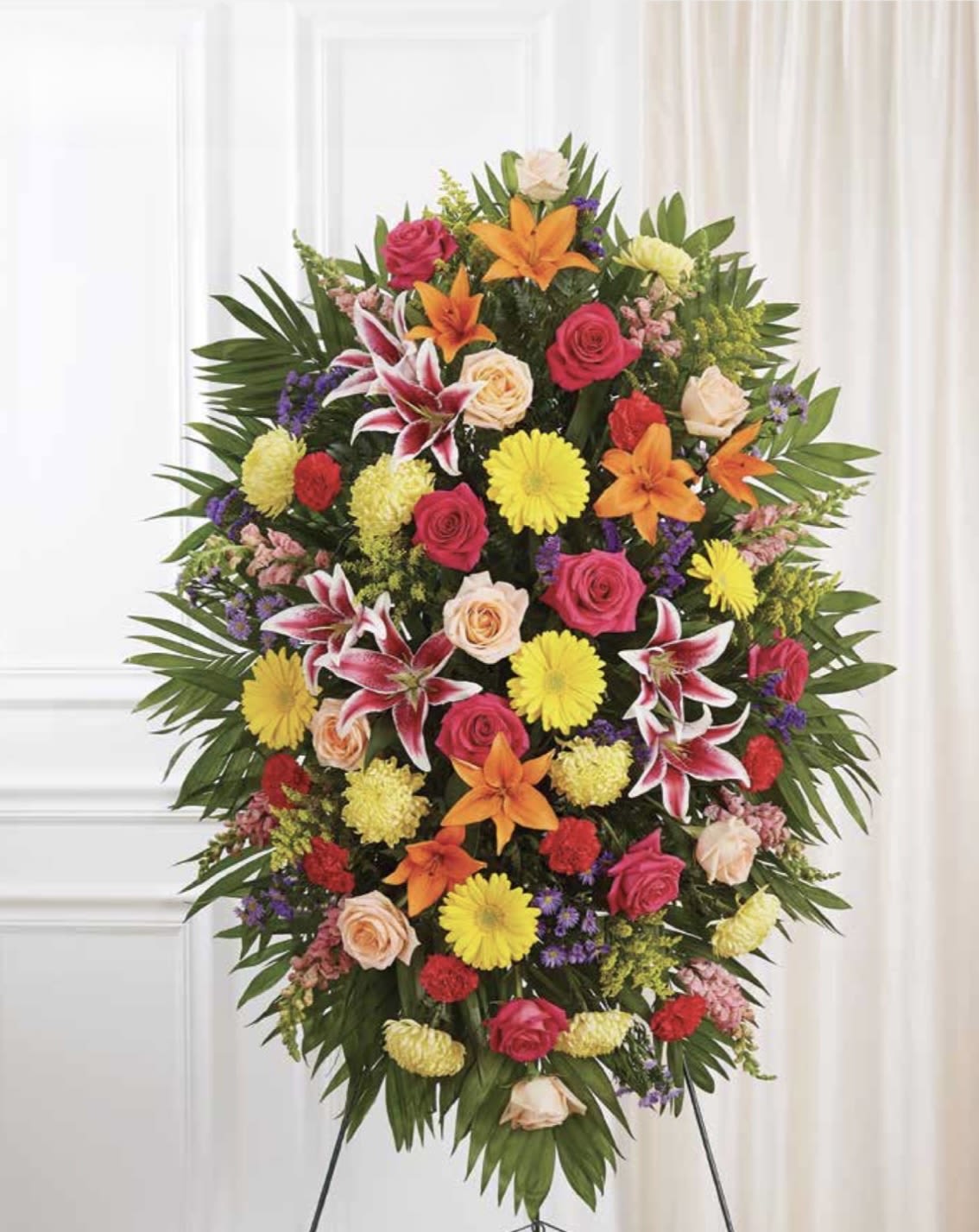 Bright Colorful Standing Spray - A vibrant standing spray that brings warmth and comfort to a sympathy or memorial setting, while still feeling celebratory and life-affirming. This bright colorful standing spray uses mixed-color blooms-pink and peach roses, orange lilies, yellow chrysanthemums, and carnations-to create a full, oval silhouette that reads from a distance and honors the person you're thinking of.  The arrangement layers large focal flowers over broad, palm-like green foliage for a formal, dense presentation on a standing easel. Textures range from the soft rose petals to the strong trumpet shape of the lilies and the cheerful pom of chrysanthemums, giving the piece visual depth and movement. Colors include mixed pinks, peaches, oranges, and sunny yellows set against deep green leaves for striking contrast.  Ideal for a funeral service, graveside tribute, or memorial celebration, this spray conveys sympathy while also celebrating a vibrant life. It arrives ready on a sturdy easel for immediate display; minimal care is needed beyond keeping it cool and out of direct sun if it will sit for extended periods before the service.