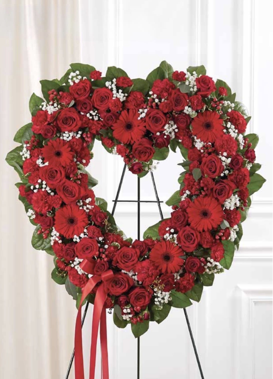 Red Open Heart - A large heart-shaped sympathy arrangement in vivid red-this floral tribute combines roses, gerbera daisies, and carnations to convey love and remembrance for a funeral or memorial service. The Red Open Heart is designed for sympathy and tribute occasions, giving the person you're honoring a bold, romantic symbol of devotion that reads clearly from a distance.  Densely arranged red roses, red gerbera daisies, and red carnations form the heart, with small white filler flowers sparking gentle contrast and broad green foliage framing the shape. A red ribbon is tied near the lower left for a polished finish, and the piece is mounted on a metal easel for easy display at a service. The symmetrical, formal construction emphasizes respect and heartfelt sentiment.  Choose this heart-shaped wreath when you want the recipient's family to feel deeply seen and honored-a meaningful way to say you're thinking of them. The arrangement arrives ready to display on the included easel; for best appearance, keep flowers cool and sheltered from direct sun until service time.