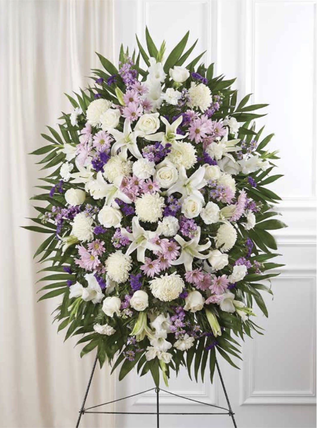 White with Lavender Standing Spray - Soft white lilies and roses set against touches of lavender and pink create a calm, dignified sympathy standing spray that honors a life with quiet grace. This white with lavender standing spray is designed as a memorial or funeral tribute-the oval, wreath-like shape and muted palette convey respect and comfort for the person you're remembering.  Dense clusters of white lilies, white roses, and white chrysanthemums are accented by small lavender- and pink daisy-like blooms and broad dark green foliage for a full, symmetrical composition. The arrangement is presented on an easel stand and framed to read clearly from front pews or beside a casket, offering visual presence without excess. The overall effect is serene and formal, the soft purples lifting the whites while the greenery provides structure and contrast.  As a sympathy piece it expresses condolences and remembrance in a tasteful, understated way-suitable for viewing, visitation, or graveside service. Please note this standing spray is intended for memorial use and arrives ready to display on its metal easel; if you have special wording or delivery timing for a service, include those details when ordering so we can help ensure it arrives as needed.