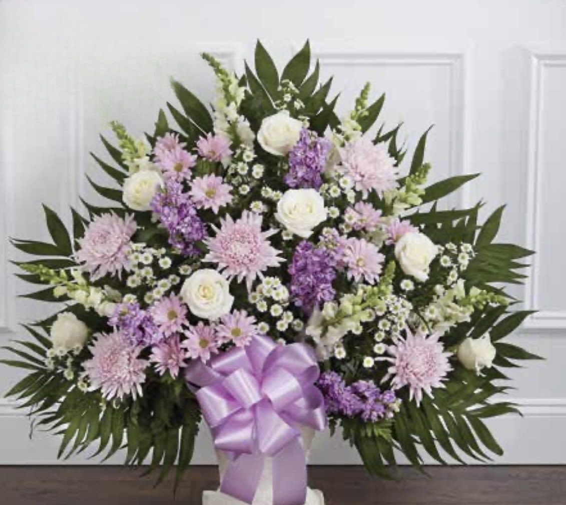White with Lavender Pedestal Arrangement - A soft, dignified arrangement that offers quiet comfort - the pedestal arrangement is a sympathy arrangement designed to express heartfelt condolences with grace. White roses and pale pink chrysanthemum-like blooms form the rounded, full silhouette while vertical snapdragon accents and clusters of small white filler flowers add gentle height and movement, making this a fitting choice for memorial services or to honor a loved one.  Deep, pointed greenery frames the arrangement for a formal, structured look, and a prominent lavender satin ribbon bow at the base provides a refined finishing touch. This piece presents beautifully in chapel aisles or beside a casket; its pastel palette of purple, white, pink, and green keeps the feeling subdued and comforting.  Choose this arrangement to convey sympathy, remembrance, or peaceful support - the kind of floral tribute that helps the person you're honoring feel respected and remembered. Arrives ready for easy placement; light misting and fresh water at the source will keep blooms looking their best through the service.