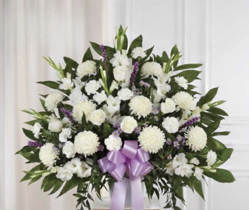 Large White with Lavender Pedestal Arrangement $325 - A broad, dignified pedestal spray crafted in white and lavender-this large white with lavender pedestal arrangement is made for sympathy and memorial services. The dense display of round white chrysanthemums and clustered white blooms, softened by pale purple accents, creates a calm, reverent presence that will help the person you're honoring feel remembered and respected.  Deep green foliage frames the symmetrical design, giving the spray a formal, full silhouette that reads beautifully from a distance. White roses and carnations blend with the chrysanthemum heads for varied texture, while a lavender satin ribbon at the base and a standing spray easel complete the presentation. Sized as a large pedestal piece, it's intended to be displayed at funerals or memorials; minimal care is required beyond keeping it cool and out of direct sun before service.