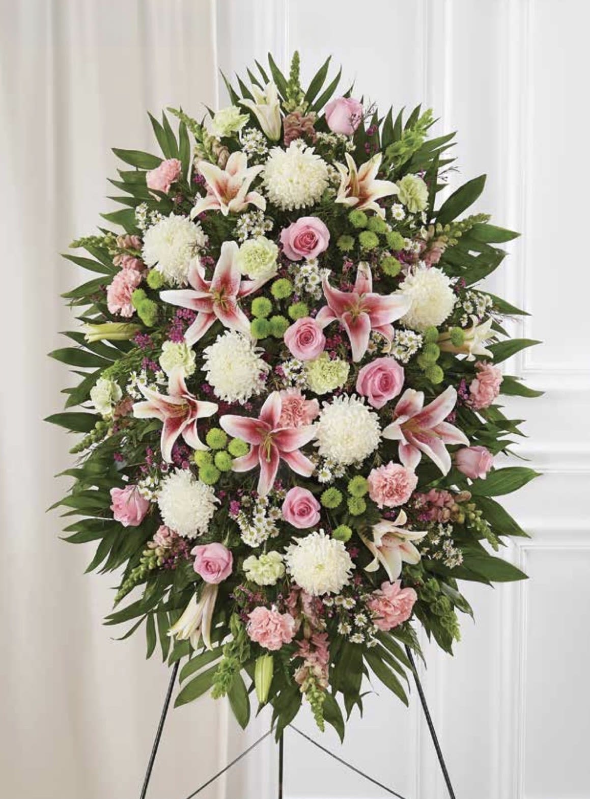 Pastel Standing Spray with Stargazer Lilies - A soft, formal tribute of pastel lilies and roses-this standing spray with stargazer lilies is designed for sympathy and memorial services. The tall oval silhouette and easel stand give it the presence and ceremony appropriate for funerals or a service remembering someone dear, offering a serene focal point that honors their memory.  Stargazer lilies take center stage amid blush roses, pink carnations, and white chrysanthemums, with touches of green foliage and deeper leaves framing the arrangement for contrast. Flowers are closely layered into a dense, symmetrical form so the color story reads softly from a distance; the easel display emphasizes the arrangement's height and traditional style.  For a comforting message at a memorial or celebration of life, this spray conveys sympathy and respect-the kind of floral piece that helps the grieving person feel seen and honored. Arrives ready on an easel stand; suitable for placement near the casket, podium, or memorial table.