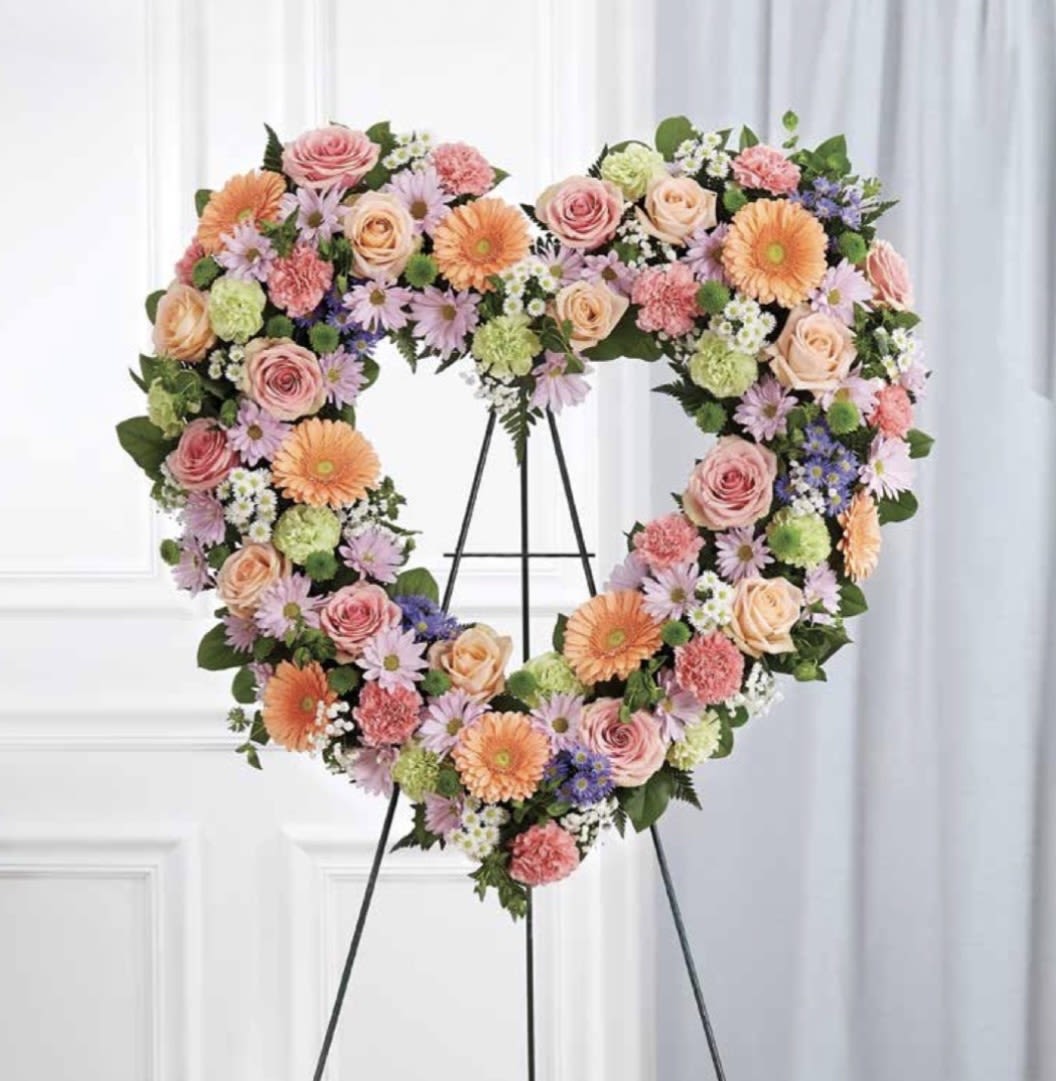 Pastel Open Heart - A large heart-shaped floral tribute that reads as tender and celebratory-perfect as a sympathy display or anniversary remembrance. The Pastel Open Heart arrangement places the heart motif and occasion in the first moment, offering the person you're honoring a gentle, comforting presence whether at a service, reception, or home tribute.  Layers of pink roses, peach gerbera daisies, lavender daisy-style blooms, carnations and small clustered fillers are arranged densely to create a polished, event-ready silhouette. Soft greens peek through the pastel palette to add depth and a natural touch. The heart is presented on a sturdy black metal easel so the design reads clearly from a distance and holds its shape throughout the day.  Choose this piece when you want the recipient to feel cherished and remembered-a statement that balances celebration and reverence. The easel-mounted format makes setup simple for venues and homes; care is routine for professional arrangements (keep out of direct heat and mist sparingly if needed).