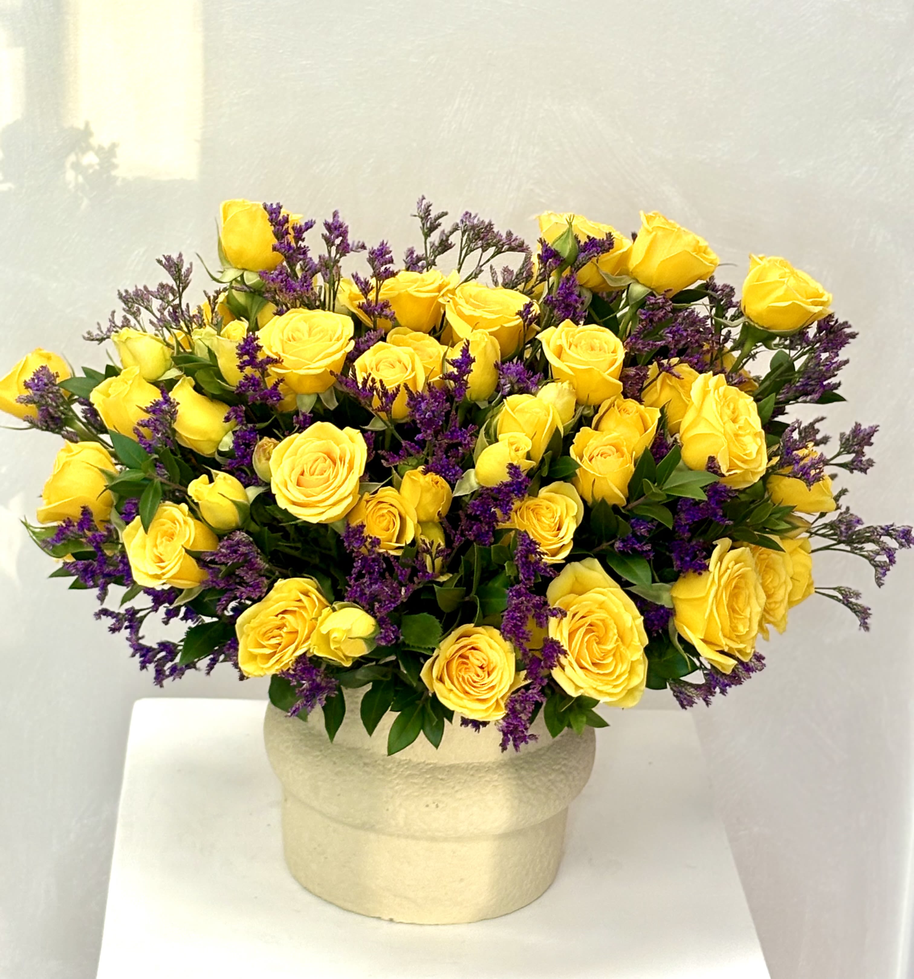 Rounded arrangement of yellow roses with purple filler flowers in a cream ceramic pot