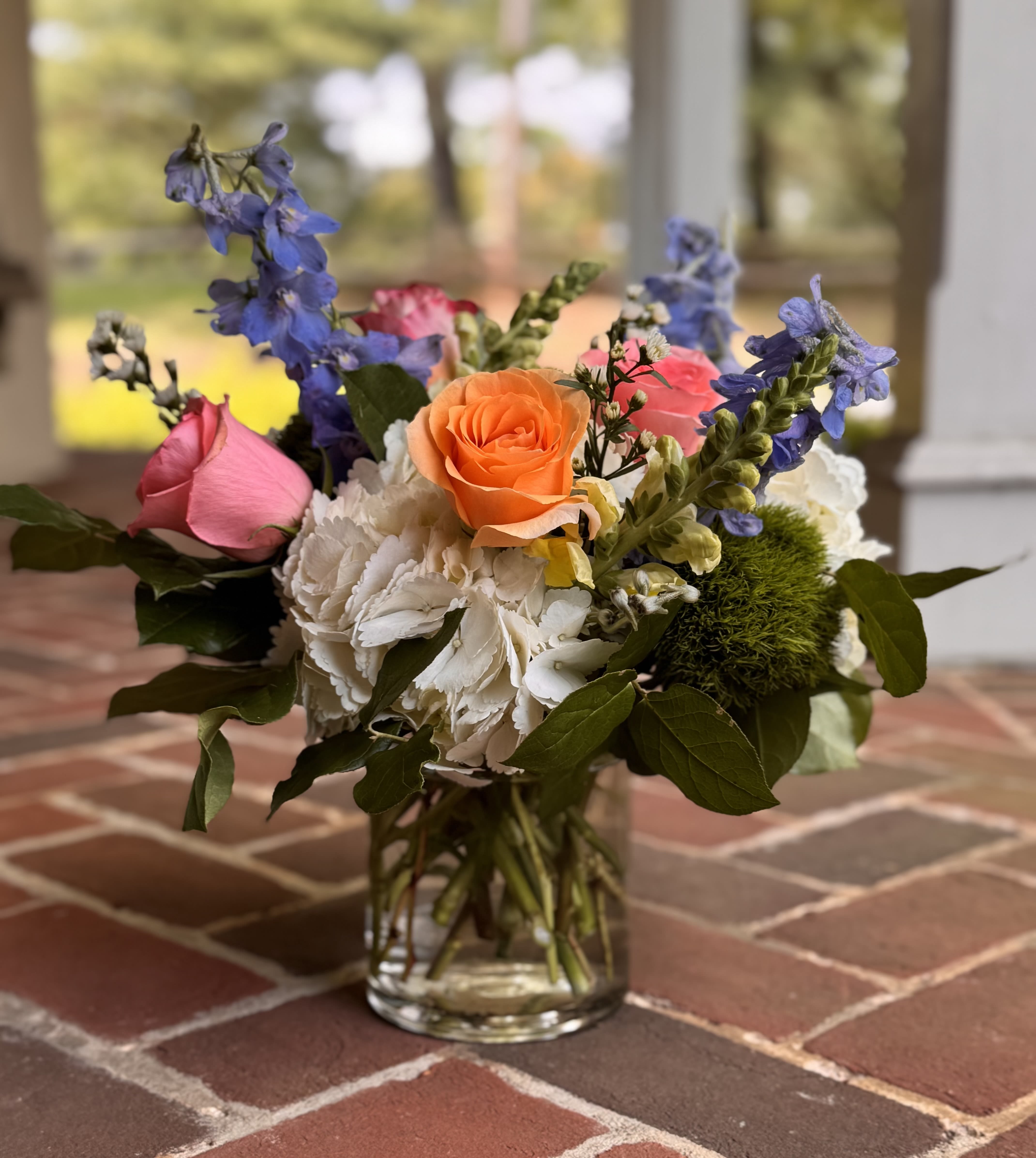 Meadow Bliss - A bright orange rose steals the show in this garden-style flower arrangement, paired with pink roses, soft white hydrangeas, and tall blue snapdragon spikes-perfect for everyday or as a birthday surprise for the person you're celebrating. The mix of rounded blooms and vertical accents creates lively, layered texture so she'll feel truly seen the moment the vase is set down.  Arranged in a clear glass vase, the palette of pink, white, blue, and orange reads cheerful and fresh, with hydrangea rounds balancing the stronger rose and snapdragon colors for a full, everyday look. The combination of roses, hydrangeas, snapdragons, and complementary stems gives a natural, springtime charm that also works beautifully as a thank-you gift or a "just because" pick-me-up.  This size sits comfortably on a mantel or dining table and arrives ready to display-simply refresh the water every few days and trim stems as they soften to extend bloom life. Whether it's for a birthday, a note of gratitude, or a seasonal celebration, this arrangement sends a warm, heartfelt message to the loved one you're thinking of.