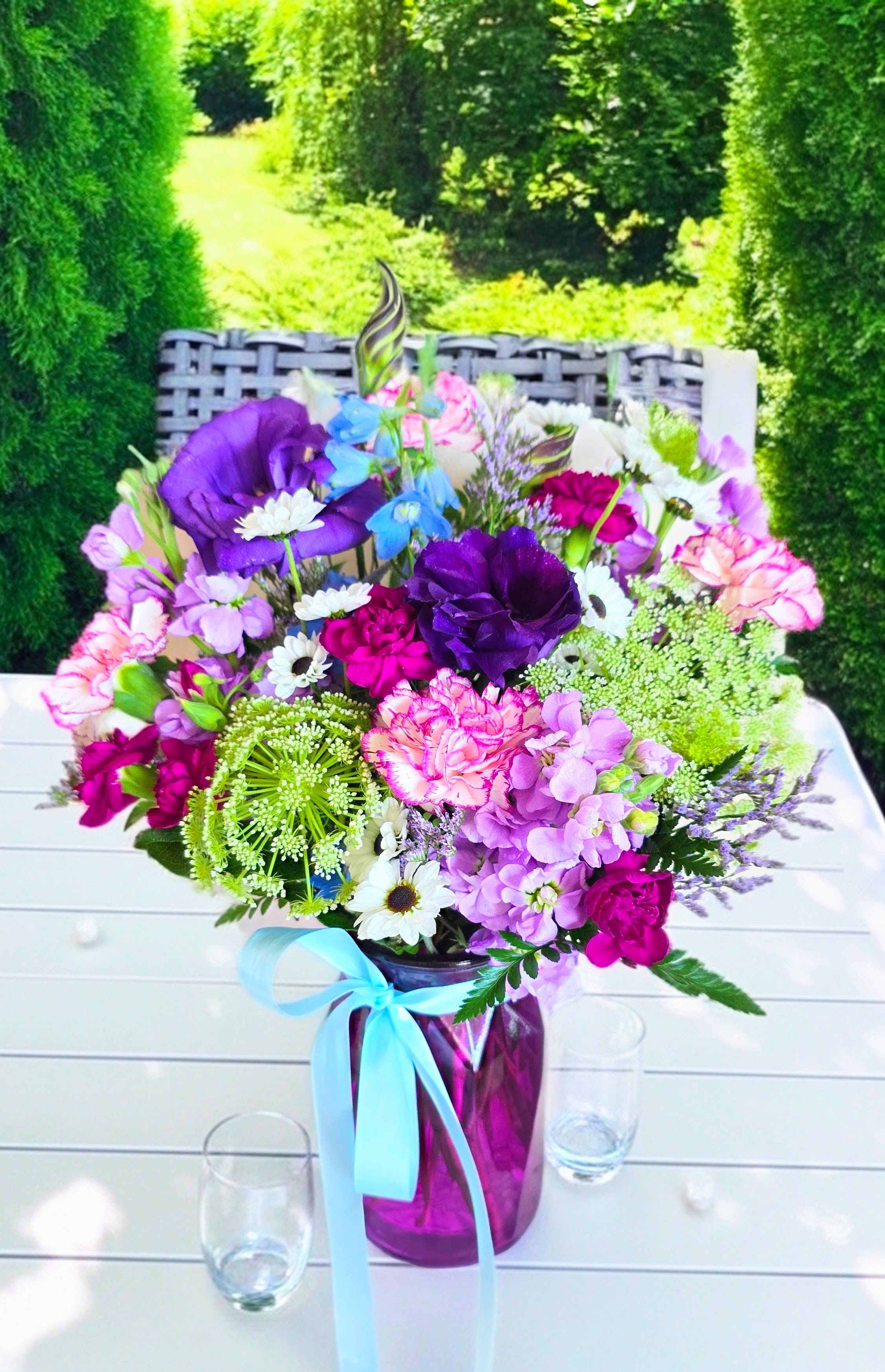 Harmony In Blossom - A lively garden-style flower arrangement that greets them with soft purples, lavender, whites and sky blues-perfect as a birthday bouquet, for him or her or a thank you gift. Gathered in a low purple glass vase and finished with a light blue satin ribbon, this composition feels polished and gift-ready keeping the arrangement easy to display on a dining table or entry console; the glass vase holds water for immediate enjoyment and simple care-freshen water every 2-3 days and trim stems slightly to keep blooms at their best.