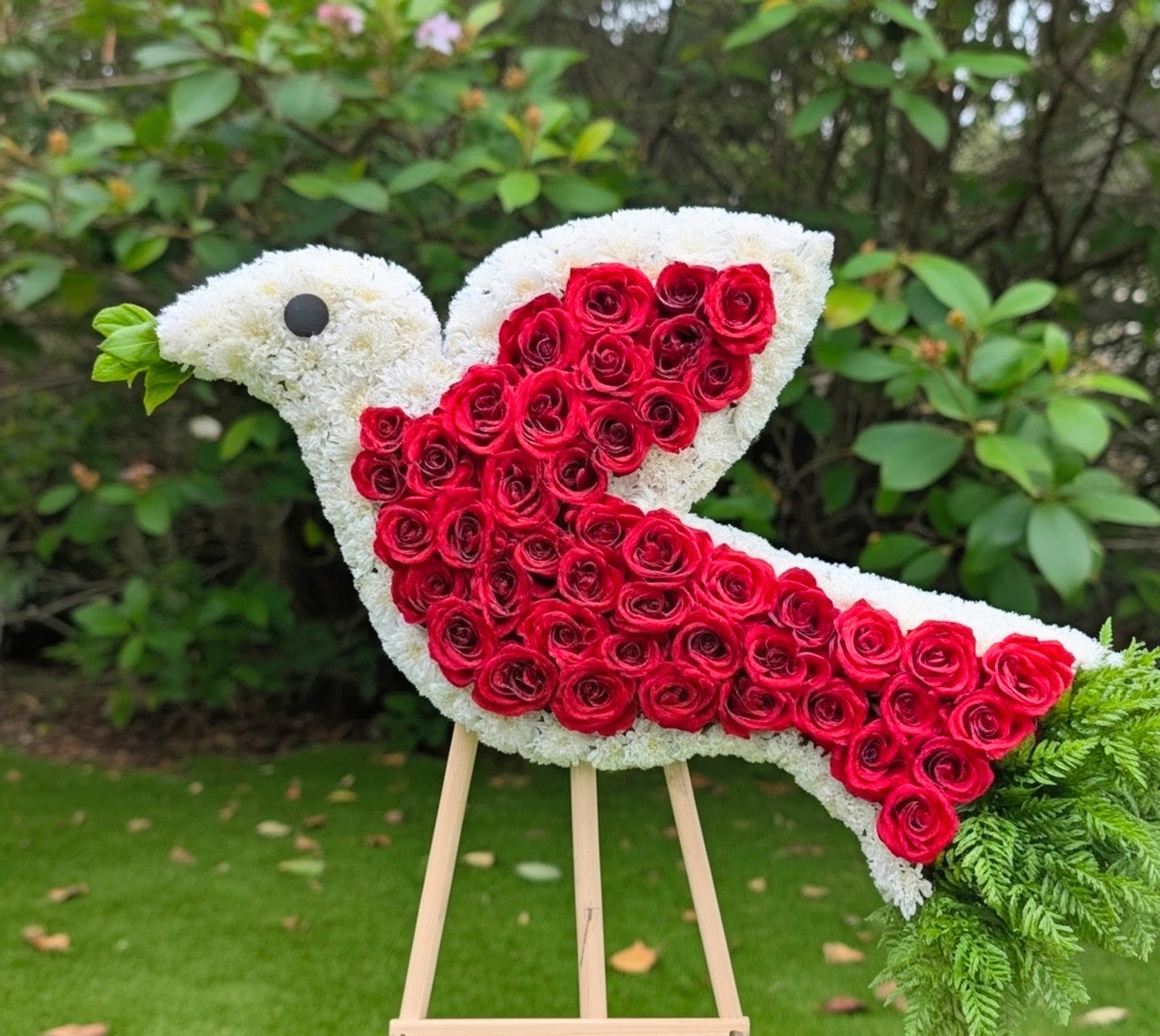 Eternal Dove Sympathy Spray - The Eternal Dove Sympathy Spray greets mourners with a sculptural white dove rendered in large red rose blooms. A sympathy arrangement designed to convey peace and loving remembrance.  Crafted on a sturdy wooden easel, the spray features a crisp white field of blooms shaped into the bird's body, a concentrated block of red roses forming the wing and tail, and fern-like greenery trailing gently from the tail for natural movement. A small dark accent marks the eye, giving the piece a thoughtful, finished look. Sized to stand prominently at a service, it's intended as a display piece rather than a handheld bouquet and will hold up well when placed at a graveside or chapel.
