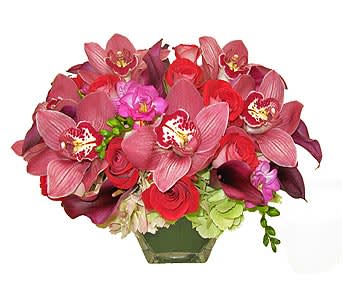 Low arrangement of pink cymbidium orchids, red roses, and dark calla lilies in a clear glass vase