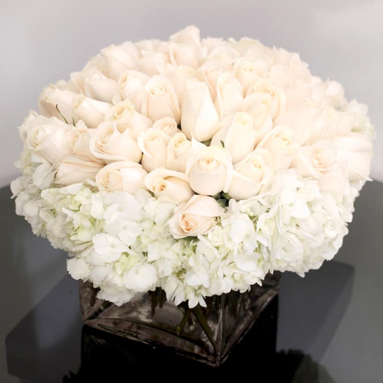 Low square vase of ivory roses surrounded by white hydrangeas in a compact arrangement