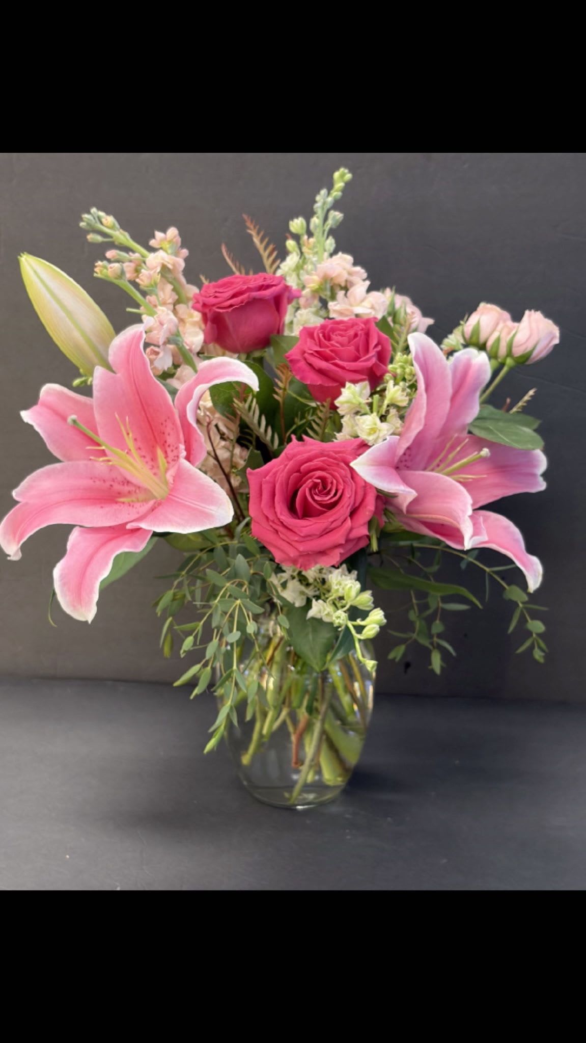 Fragrant Vase of Lilies and Roses - This arrangement pairs large pink lilies with hot pink roses, pale white and blush stock and filler blooms, plus soft greenery spilling around a clear glass vase for a classic, unfussy presentation. Colors include pink, white, yellow, lavender and mixed tones that catch the eye without overpowering. Sized to sit on a bedside table or dining table, the vase and stems are arranged for lasting structure - trim stems and refresh the water every few days to keep blooms at their best.  Ideal for Mother's Day, Valentine's Day, anniversaries, birthdays, or simply to say "get well soon" or "I'm thinking of you," this bouquet sends a warm, personal message that will make the recipient feel genuinely seen and cared for. Order for same-day local delivery when available to brighten their day quickly.