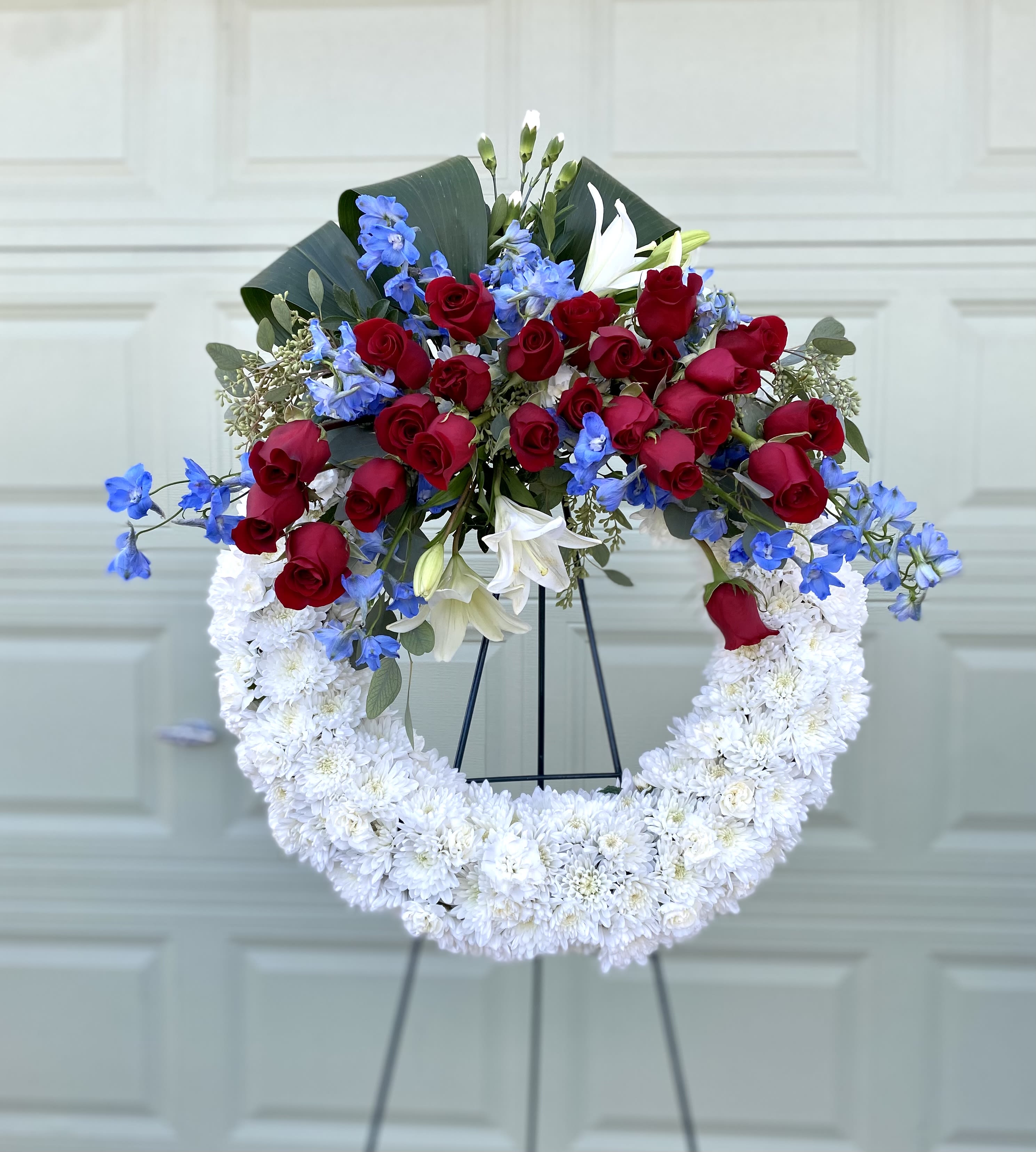 Red roses, blue flowers, and white lilies in a white chrysanthemum wreath