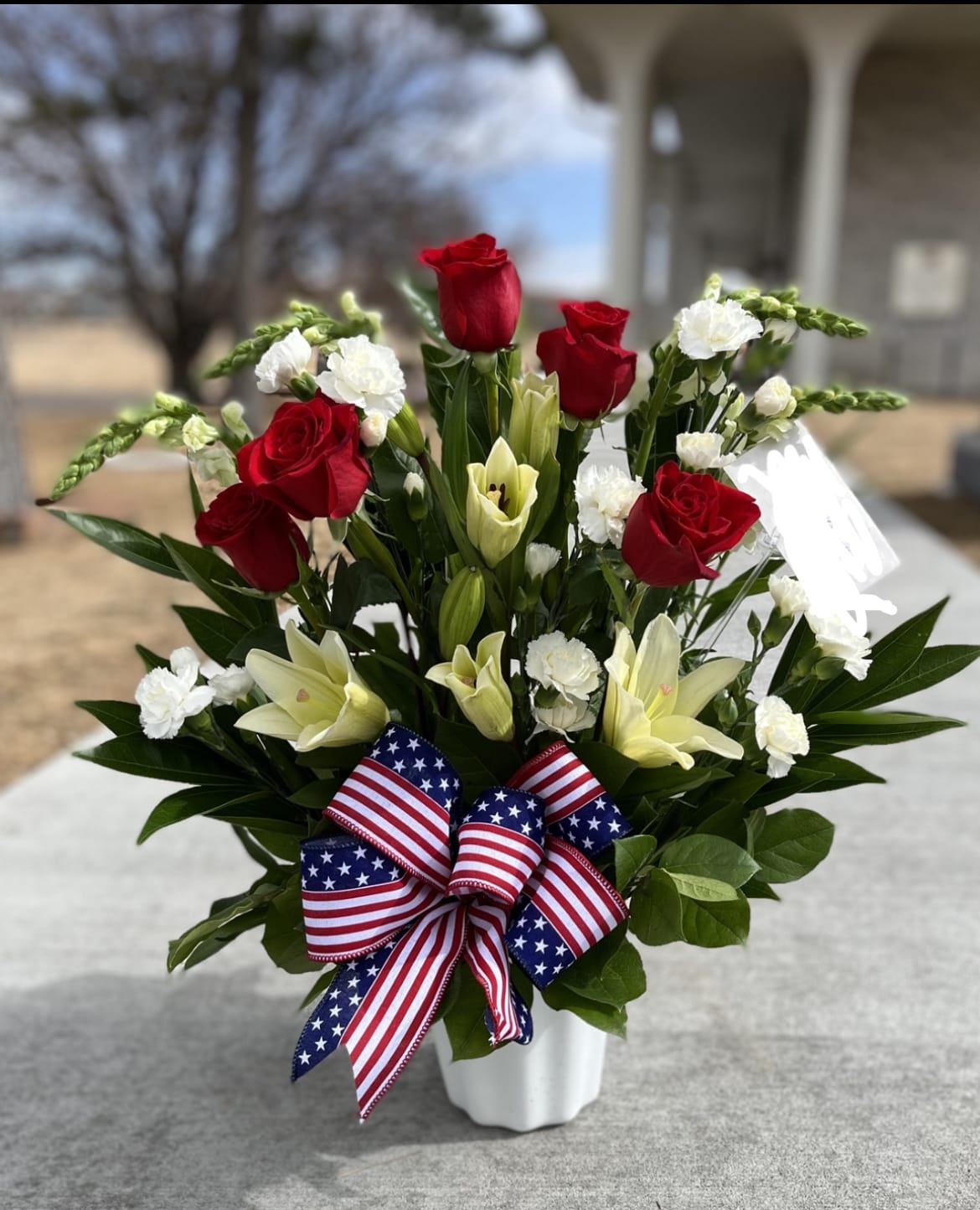 Red roses and white lilies in a white vase with an American flag ribbon
