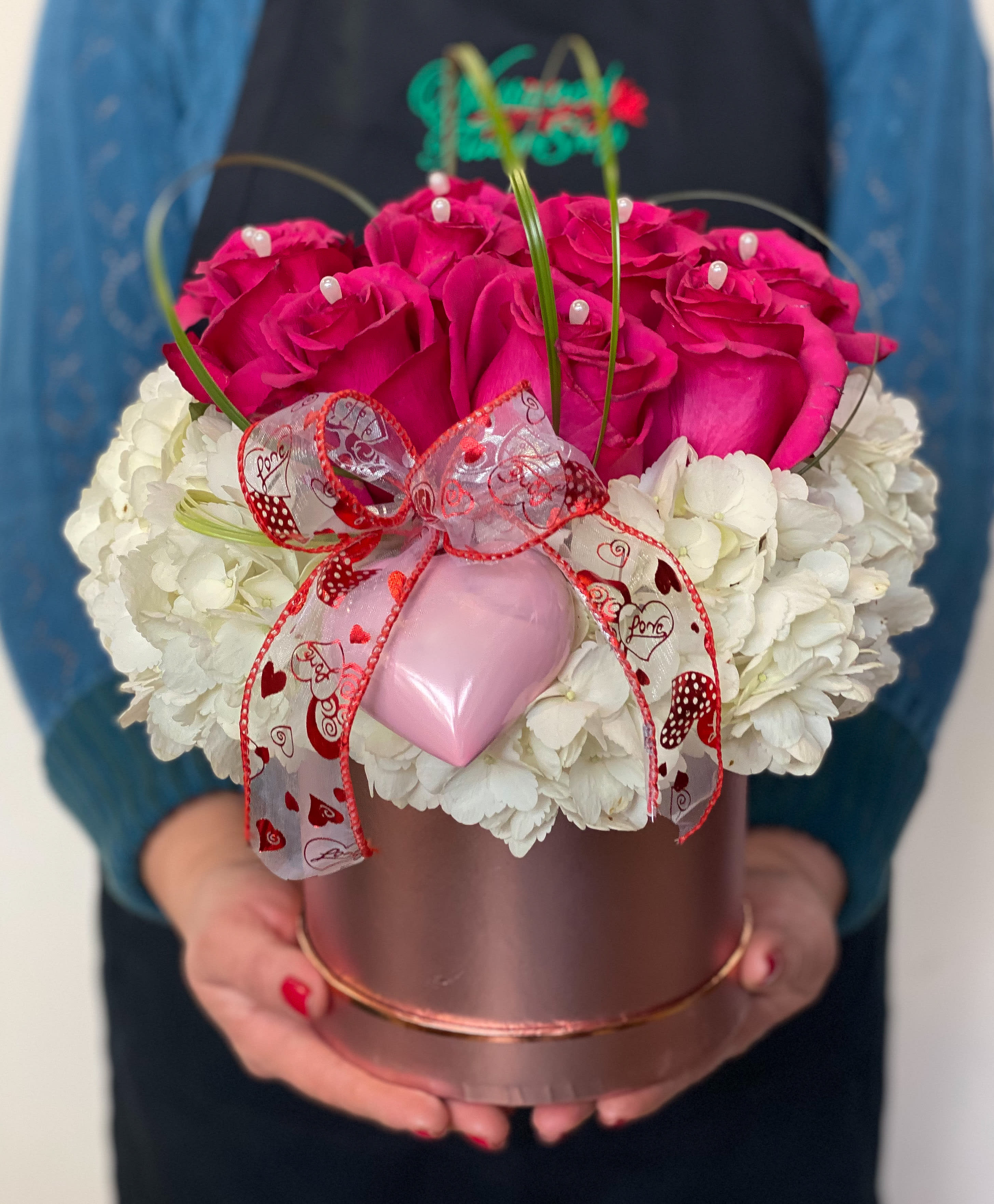 Pink roses and white flowers arranged in a round gift box with a ribbon and heart ornament.
