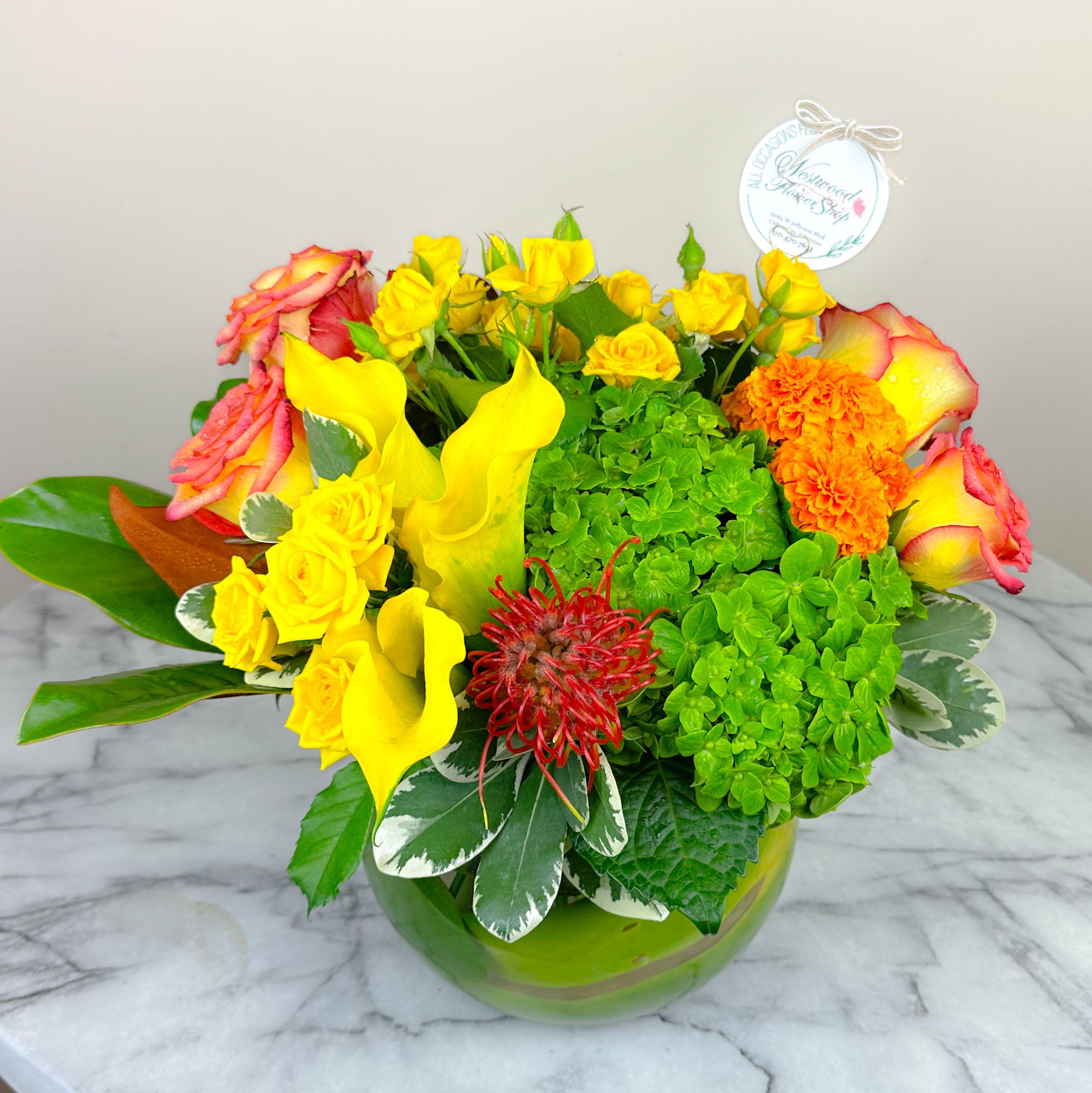Bright mixed bouquet with yellow roses, calla lilies, and orange blooms in a green glass vase