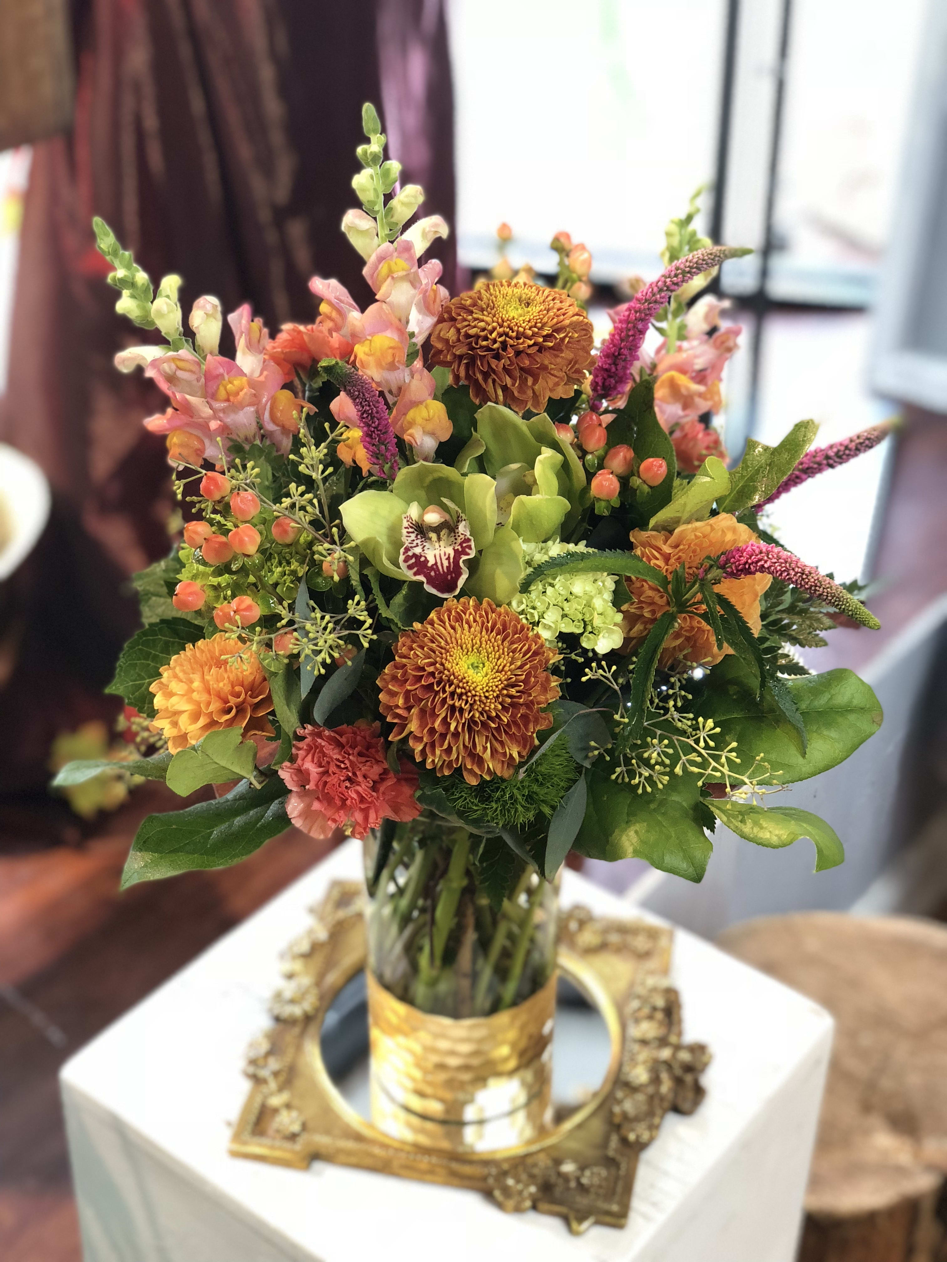 Tall mixed arrangement with orange mums, snapdragons, and green orchid in a glass vase with gold band
