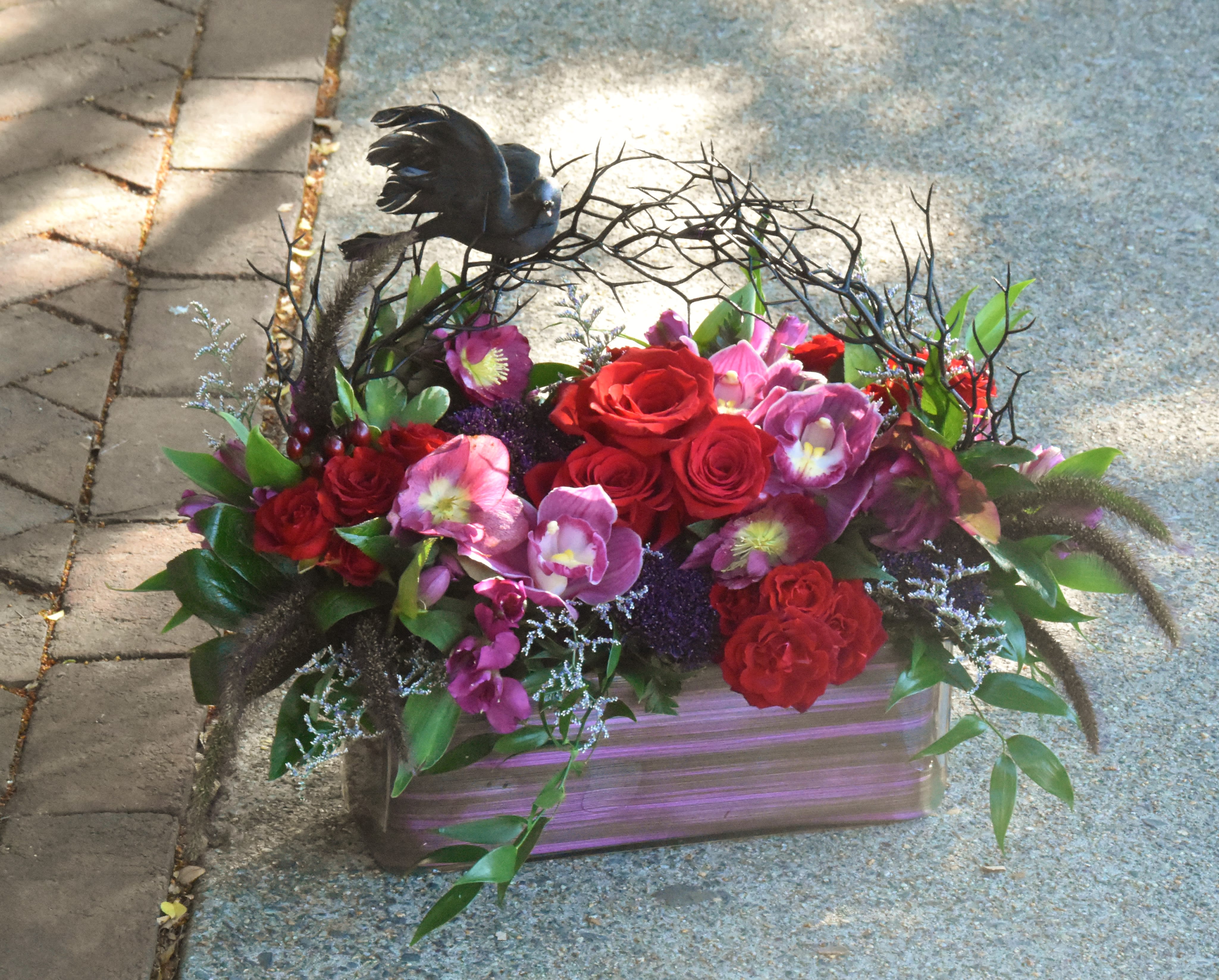Low rectangular arrangement of red roses and purple flowers with a black bird on twig accents