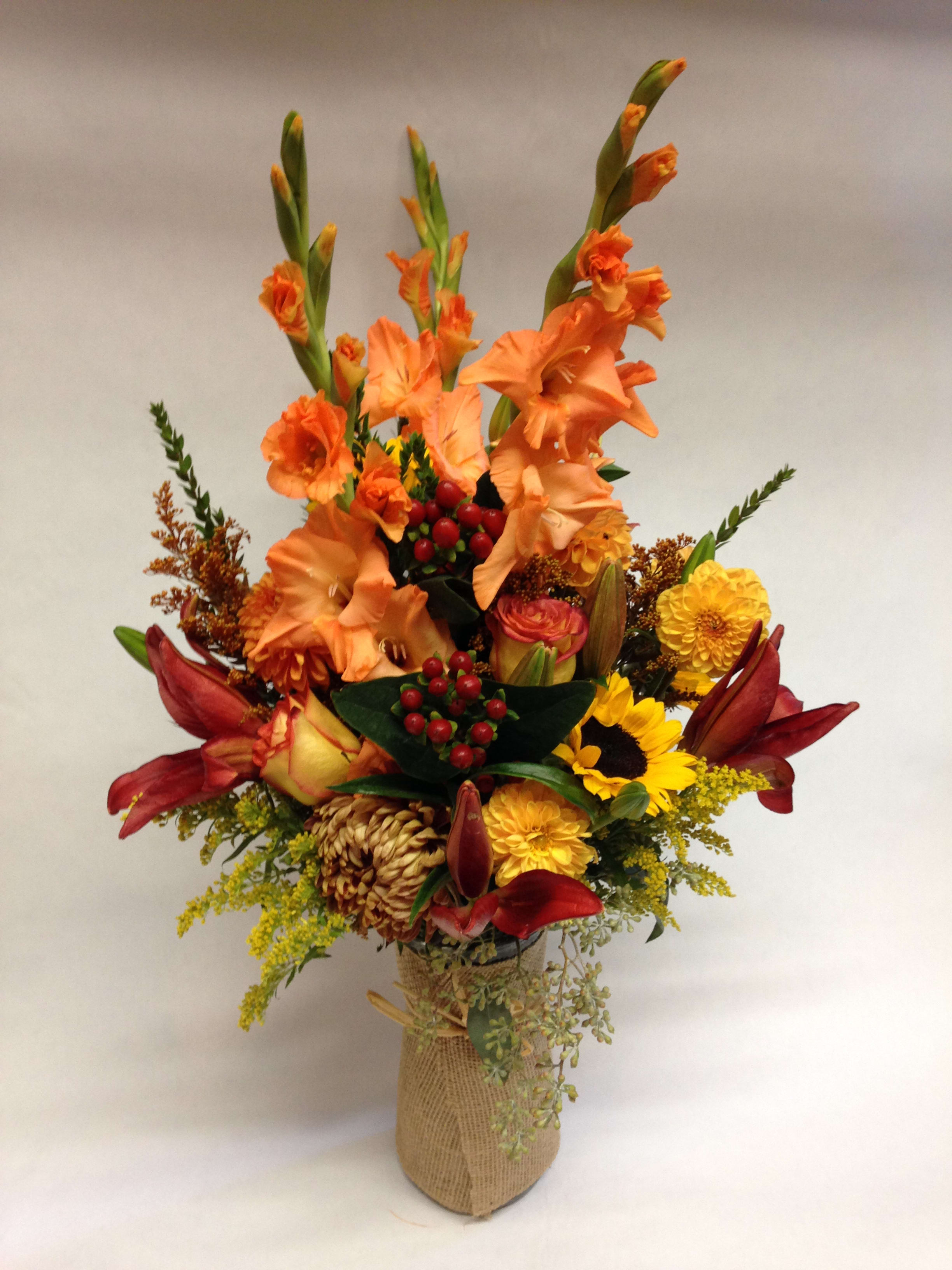 Tall mixed bouquet with orange gladiolus, red lilies, and yellow blooms in a burlap-wrapped vase