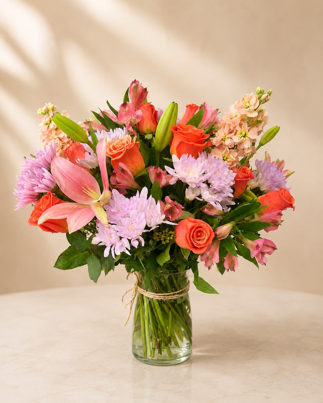 Coral Kisses - Description &amp; Substitution Policy  Brighten someone’s day with the warmth and beauty of Coral Kisses. This cheerful garden-style arrangement features vibrant coral roses, graceful pink lilies, soft lavender cushion chrysanthemums, peach stock, and delicate pink alstroemeria, beautifully designed in a classic clear glass vase. Its fresh, elegant color palette makes it a wonderful gift for birthdays, anniversaries, congratulations, thank-you gifts, Mother’s Day, or simply to let someone know you’re thinking of them.  Our expert designers handcraft each arrangement using the freshest premium flowers available. Due to seasonal and regional availability, some flowers or the vase may be substituted with blooms or containers of equal or greater value while maintaining the overall color palette, style, size, and premium quality of the arrangement. Every design is created with care to ensure it arrives just as beautiful as the one pictured.