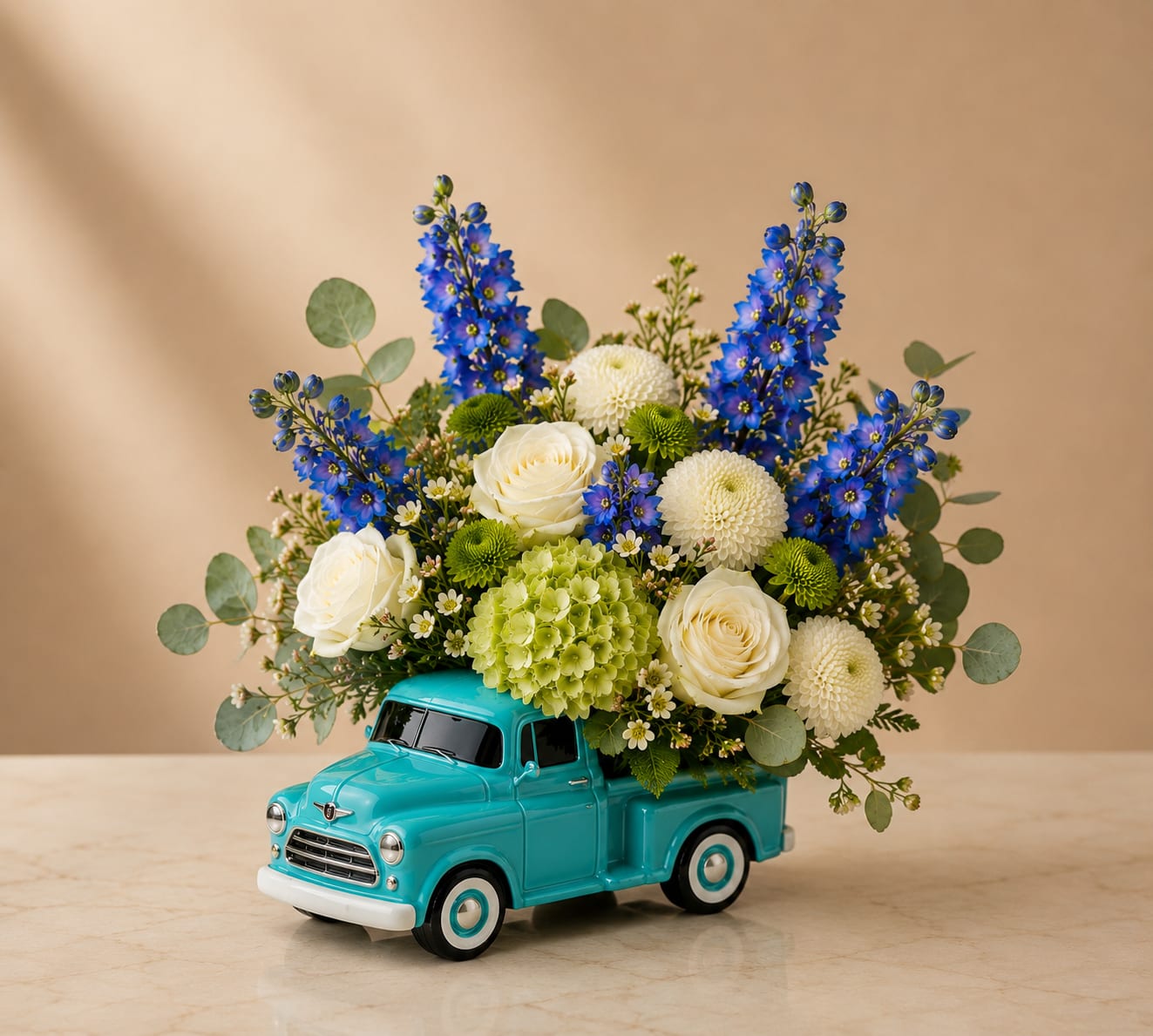 Country Blue Bloom Truck - Inspired by open roads, hard work, and timeless memories, Dad’s Country Drive is a classic Father’s Day arrangement designed in a charming vintage pickup truck keepsake vase. Featuring vibrant blue delphinium, elegant white roses, green hydrangea, white button poms, green button poms, wax flower, and eucalyptus, this arrangement offers a fresh and sophisticated look with a touch of rustic charm.  The blue and white color palette symbolizes loyalty, strength, trust, and appreciation, making it a meaningful gift for fathers, grandfathers, husbands, and father figures alike. The collectible truck vase serves as a lasting reminder of your thoughtful gesture long after the flowers have been enjoyed.