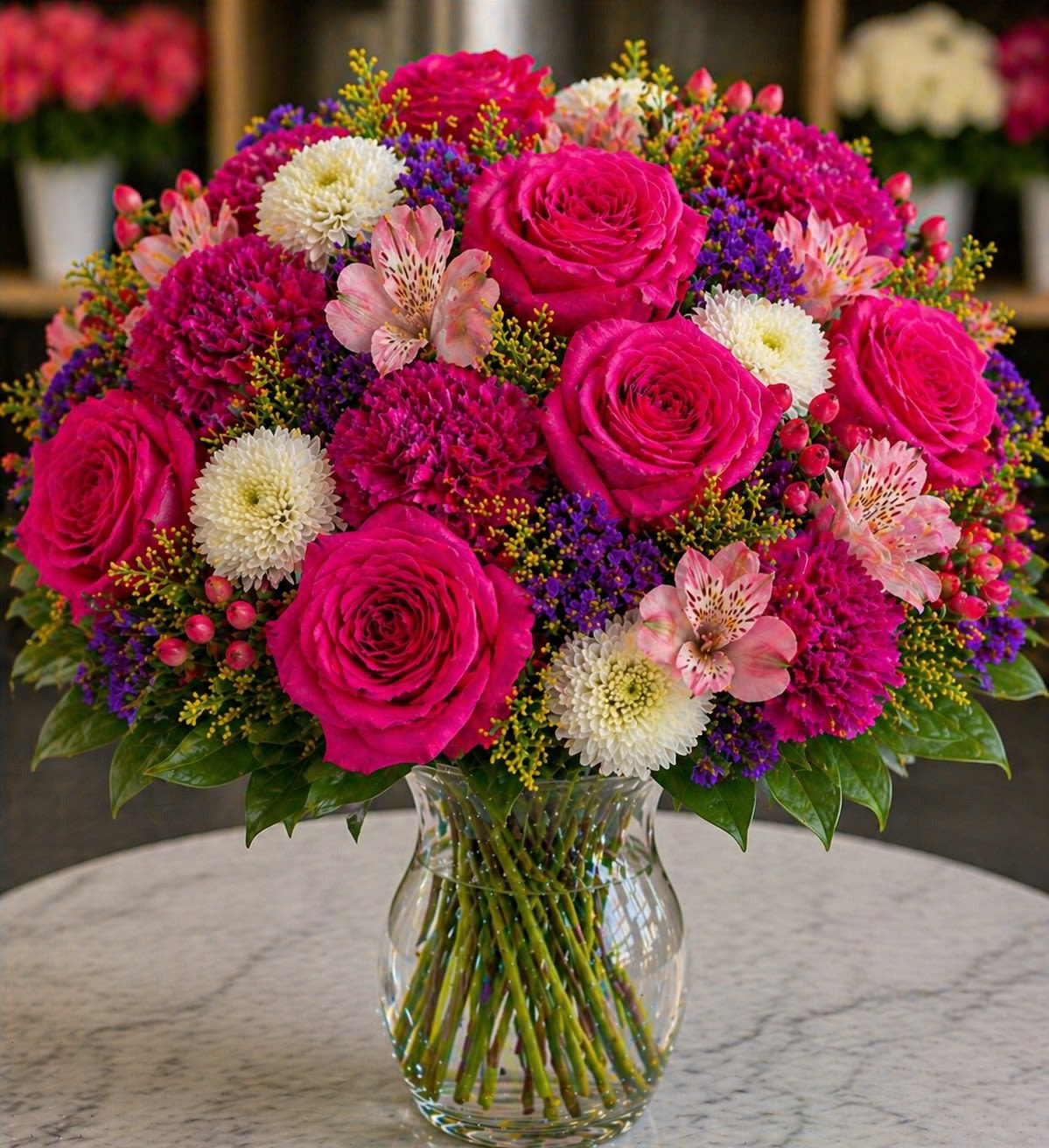 Fuchsia Bellezza - A vibrant luxury floral arrangement featuring bold fuchsia roses, rich magenta carnations, delicate blush alstroemeria, crisp white button mums, vivid purple statice, golden solidago, and seasonal pink hypericum berries in a classic clear glass vase. Fuchsia Bellezza radiates glamour, femininity, and modern elegance. The striking jewel-toned pinks paired with soft blush accents and deep violet textures create an unforgettable bouquet full of energy and sophistication.
