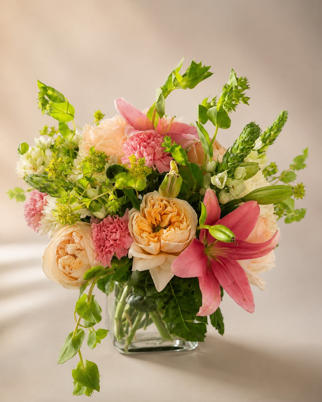 Garden Melody - Description &amp; Substitution Policy  Inspired by the beauty of an English garden, Garden Melody is a lush, elegant arrangement that brings together soft textures, romantic blooms, and fresh seasonal greenery. Featuring premium garden roses, fragrant pink lilies, soft pink carnations, white cushion chrysanthemums, white stock, Bells of Ireland, and lush foliage, this stunning design is artistically arranged in a modern clear glass cube vase. Its graceful blend of blush pinks, creamy peaches, fresh greens, and crisp whites creates a timeless gift that’s perfect for birthdays, anniversaries, Mother’s Day, congratulations, thank-you gifts, housewarmings, or simply to brighten someone’s day.  Each arrangement is handcrafted by the designers at House of Bloom using the freshest premium flowers available. Due to seasonal and regional availability, flower varieties, colors, or the vase may be substituted with blooms or containers of equal or greater value while maintaining the overall style, color palette, size, and premium quality of the arrangement. Every design is carefully created to ensure it arrives just as beautiful as the one pictured.