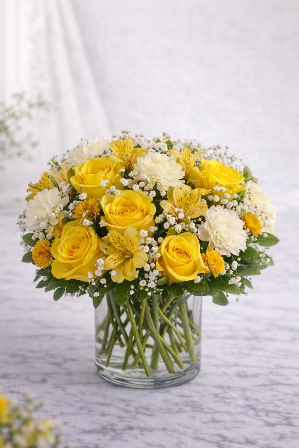 Golden Glow Bouquet -  Description:  A radiant mix of vibrant yellow roses, sunny alstroemeria, delicate white carnations, and airy baby’s breath, beautifully arranged in a clear glass cylinder vase.  This cheerful design blends bright golden tones with soft white accents and fresh greenery, creating a warm and uplifting look. The rounded, full shape makes it perfect for celebrating joyful moments or sending a burst of happiness to someone special.  Ideal for birthdays, congratulations, thank you gifts, or simply brightening someone’s day.   Substitution Policy:  Due to seasonal availability and market conditions, specific flower varieties may vary. If substitutions are necessary, blooms will be replaced with flowers of equal or greater value while maintaining the same vibrant yellow and white color palette.  The overall size, fullness, and sunny aesthetic of the arrangement will always remain consistent with the design shown.  ? Container Notice:  Vase style may vary slightly based on availability, but it will always complement the yellow and white tones of the arrangement. The overall design and color harmony will remain true to the original look.