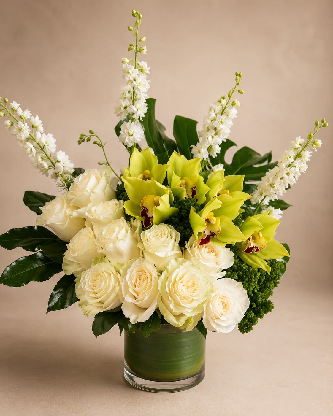 Ivory Orchid Elegance - A refined and luxurious arrangement featuring premium green cymbidium orchids, crisp white roses, delicate dendrobium orchids, and modern tropical foliage. Designed in a sleek glass cylinder with layered greenery, this piece blends soft ivory tones with rich emerald accents for a timeless and sophisticated look. Perfect for anniversaries, sympathy gestures, birthdays, corporate gifting, or elegant home décor. • Premium green cymbidium orchids • Fresh white roses • White dendrobium orchids • Textural green accents • Modern glass vase included This arrangement delivers a clean, upscale aesthetic with balanced height and fullness. ⸻  Seasonal &amp; Freshness Guarantee We carefully select the freshest blooms available to ensure exceptional quality and longevity. Due to seasonal availability, certain flowers or greens may occasionally be substituted. If a substitution is needed, we will: • Maintain the same elegant white and green color palette • Preserve the modern, luxury design style • Ensure the arrangement remains full and balanced • Use flowers of equal or greater value and quality Your order will always reflect the same sophisticated look and feel shown — never a different style or theme. Each arrangement is handcrafted with attention to detail to ensure beauty and consistency.
