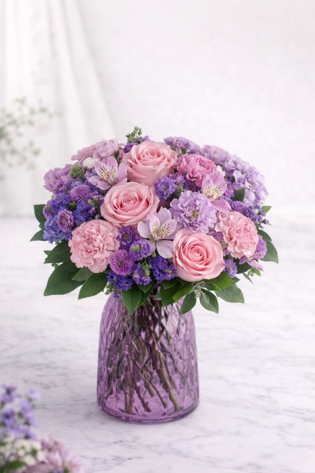Lavender Blush Elegance - A soft and romantic arrangement featuring blush pink roses, delicate lavender blooms, light pink carnations, and lush greenery, beautifully designed in a textured purple glass vase. This design blends gentle shades of pink and lavender to create a calm, graceful, and timeless look. Perfect for birthdays, anniversaries, sympathy, or simply sending love in the most elegant way.  Substitution Policy: Flower varieties may vary based on seasonal availability, but the overall color palette of soft pinks and lavender tones will always remain consistent. Any substitutions will be of equal or greater value and carefully selected to maintain the same full, romantic style shown.  Container Notice: The vase may vary slightly depending on availability, but it will always complement the arrangement and match the soft pink and lavender aesthetic. The overall look and feel will remain true to the design pictured. Our goal is to deliver an arrangement that reflects the same beauty, fullness, and color harmony as shown.