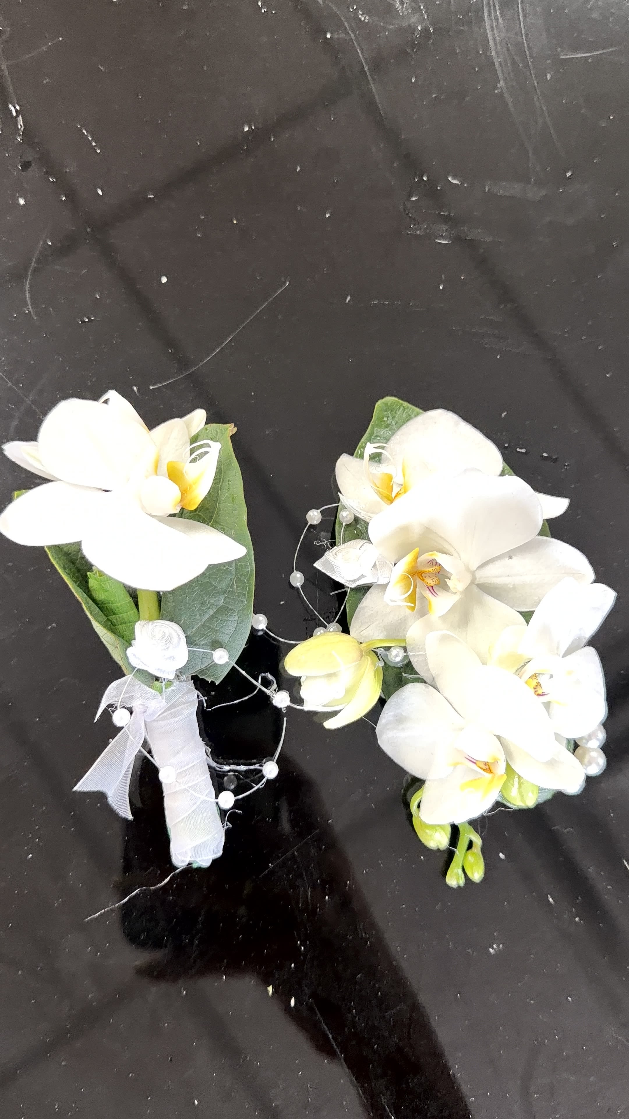 Orchids Corsage and Botonier - Cascading white Phalaenopsis orchids with pale yellow centers create a quietly striking corsage and boutonniere set perfect for wedding attire or other formal occasions. The white-and-green palette reads fresh and elegant so the person you give this to will feel graceful and noticed-an ideal choice for brides, mothers, or honored guests who deserve a refined finishing touch.  Each piece is crafted with single white Phalaenopsis blooms backed by glossy green leaves, stems wrapped in white ribbon and secured with floral tape. Decorative pearl wire and pearl picks add a soft, luminous detail while a small ribbon bow on the corsage gives a polished, wearable look. Sizes are intimate and wearable for lapels or wrist placement; handle gently and keep cool and shaded until wear to preserve the delicate orchid petals.