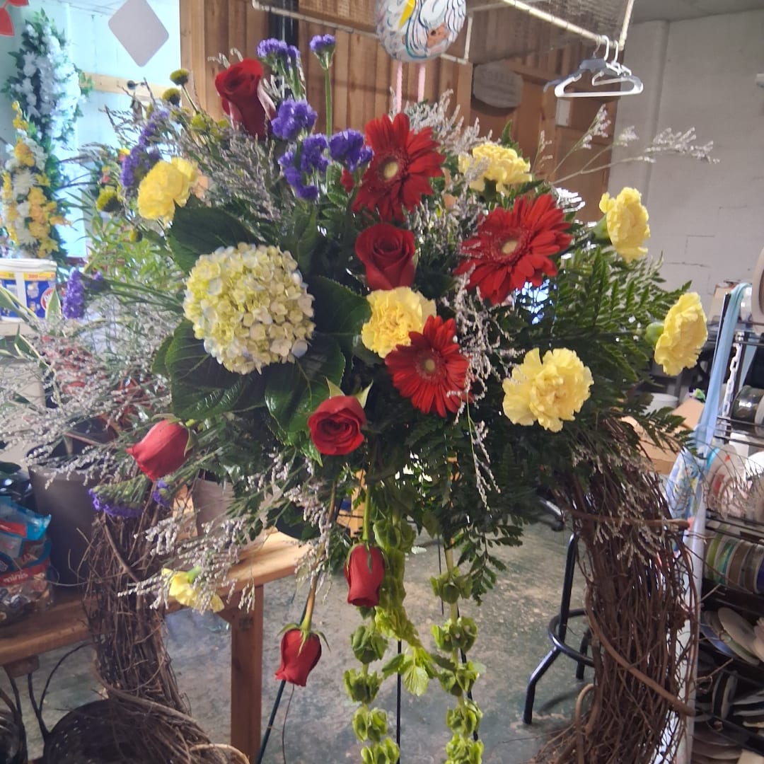 Large mixed floral arrangement with red roses, red gerberas, yellow carnations, and purple accents.