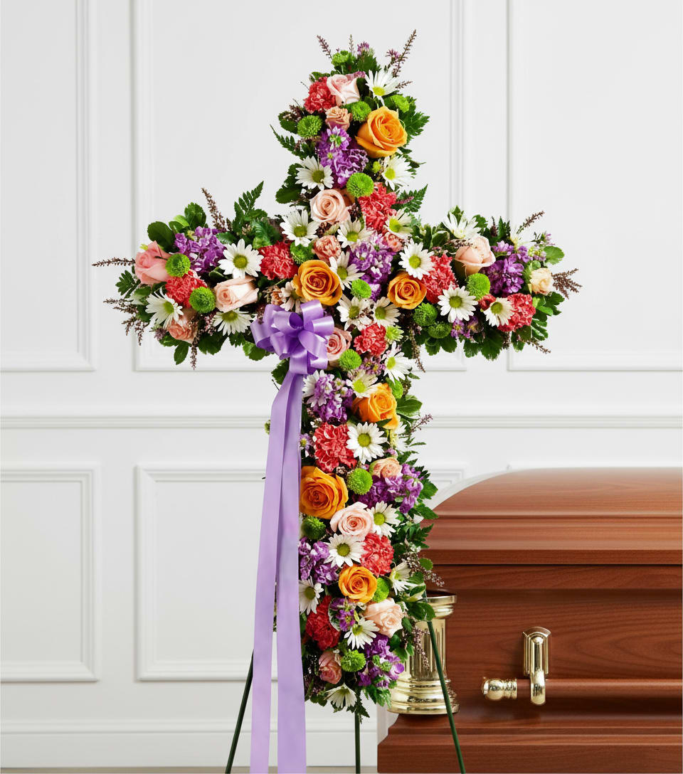 Joyful Pastel Standing Cross - A densely covered standing cross in mixed pastel and bright tones creates a comforting, reverent focal point for a funeral or memorial service. This sympathy standing cross uses roses, carnations, daisies and small accent blooms in white, pink, purple, orange and green to form a layered, textured surface; a long lavender/purple ribbon at the center adds a devotional, formal touch that draws the eye along the vertical line of the design.  Designed to mount on a metal standing easel and shown beside a casket, the cross reads clearly from a distance while offering subtle details up close - blush and orange roses, red carnations, white daisy-like blooms, purple accents and bright green button-style flowers combine for both visual warmth and solemn dignity. The arrangement's generous scale and dense flower coverage ensure it stands as a meaningful tribute that will help the person you're honoring feel remembered and seen.  Ideal for a funeral, memorial or graveside service, this standing cross conveys sympathy and respect in a traditional, ceremonial way. Available to place on a standing or casket easel; please indicate delivery details and service time at checkout so we can coordinate placement for the service.