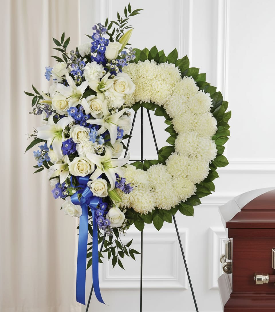 Heavenly Hearts Standing Wreath - A circular wreath of pure white chrysanthemums edged with deep green foliage creates an immediate impression of quiet dignity - a standing funeral wreath designed to honor a life with grace. This sympathy arrangement pairs classic white roses and fragrant white lilies clustered on one side with small blue accent flowers for a cool, calming contrast; the bright blue ribbon draped from the lower section adds a formal, ceremonial touch that reads respectful and intentional.  Hand-crafted on a sturdy floral wreath stand, the composition balances dense white blooms with the texture of foliage to present a clean, solemn silhouette suitable for memorial services or graveside tribute. Sized as a large standing wreath, it commands presence without overwhelming the space; the white-and-blue palette conveys compassion and peace so the person you're remembering feels honored and seen.  Choose this wreath to express condolences at a funeral or to send as a memorial tribute that speaks of reverence and care. Our florists prepare each piece to arrive fresh and secure on its floral wreath stand; for delivery timing and placement assistance, please contact us when ordering so we can ensure respectful presentation at the service.