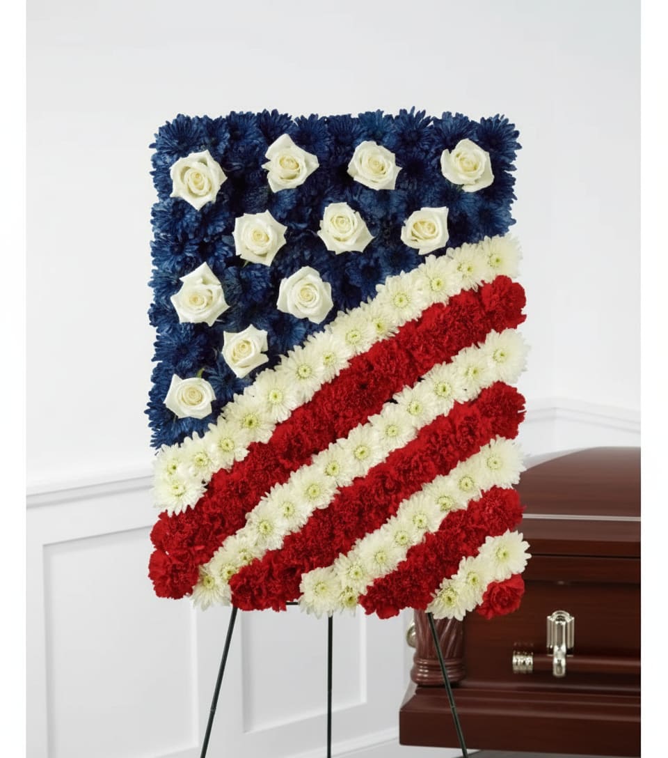 Glory Be Flag Tribute - A formal American flag tribute crafted for memorial and funeral services, this flag-shaped flower arrangement is a respectful choice for honoring military service or a patriotic life. The design reads immediately as a flag with a rich blue field accented by white roses and bold red-and-white diagonal stripes, making it a fitting sympathy tribute for a funeral or graveside ceremony.  The composition uses roses, carnations, and chrysanthemums to build a dense, textured surface that holds its shape on an easel or beside a casket. Deep blue blooms form the canton while clusters of white roses punctuate the stars area; alternating bands of red and white carnations and chrysanthemums create striking contrast and a ceremonial, sculpted profile. Sized to display on an easel, the arrangement carries a solemn presence appropriate for family, veteran groups, or official memorials.  This piece communicates honor and remembrance-the kind of tribute that helps the person you're remembering feel seen and respected. Designed for use at services or as a casket spray alternative on an easel, it arrives ready to display; standard floral-care practices (cool, sheltered display away from direct sun) will help the blooms maintain their form through the service.