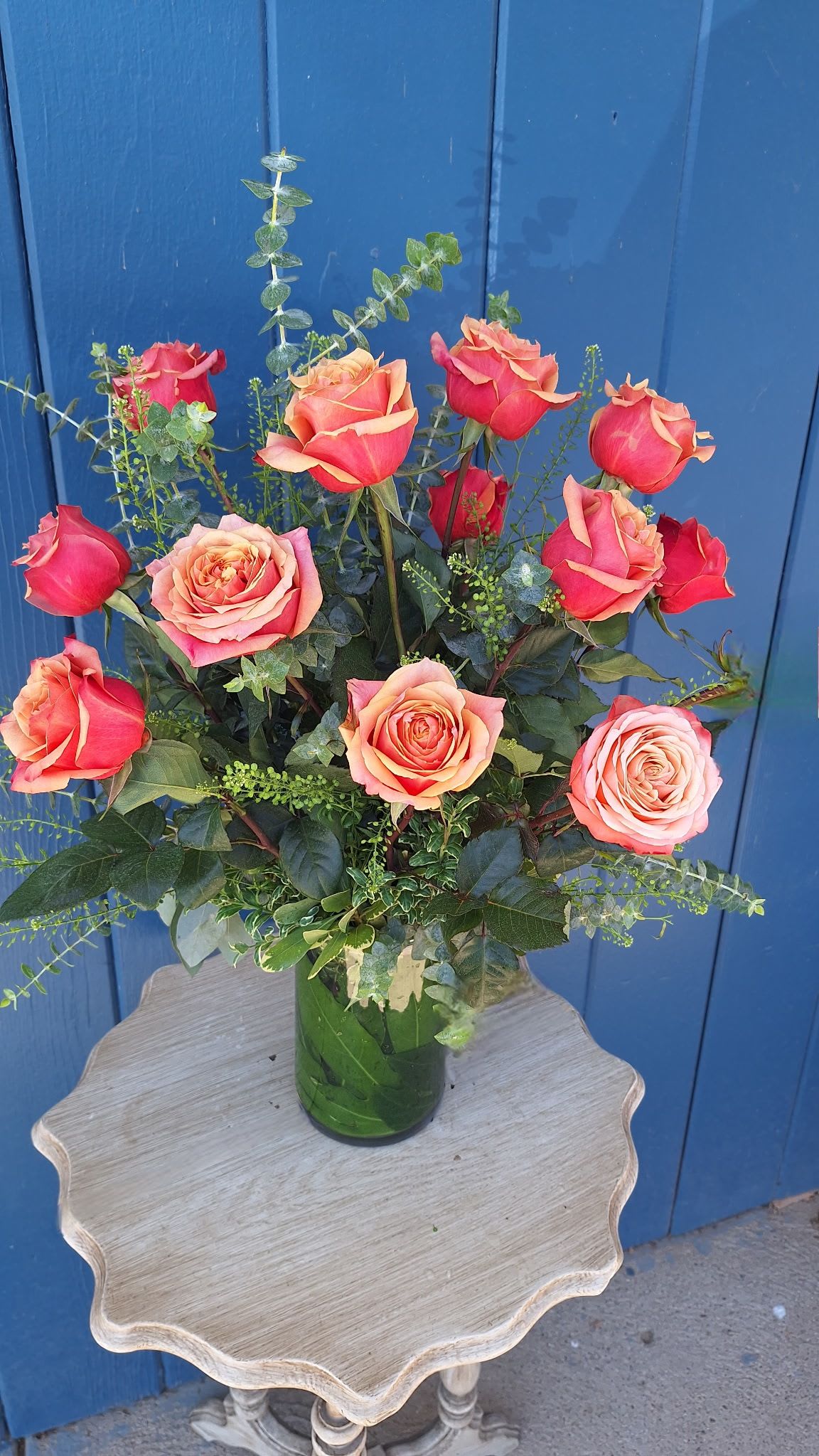 Tall arrangement of coral and orange roses in a clear glass vase on a small wooden table