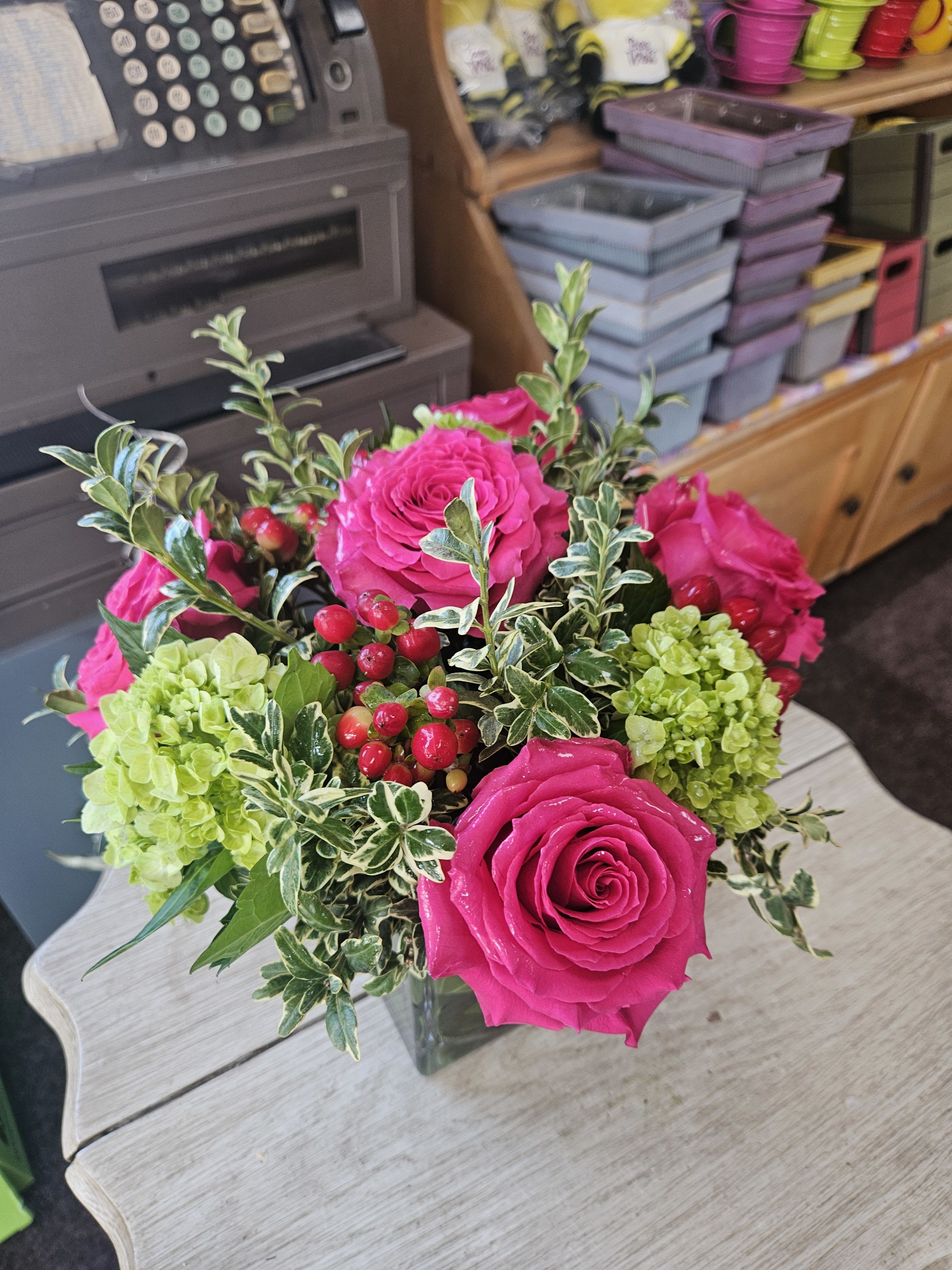 Cube vase of hot pink roses, green hydrangeas, and red berries on a light wood table