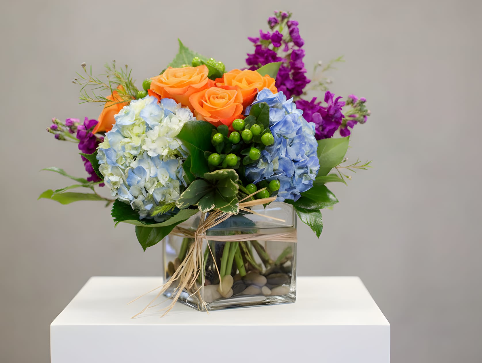 Bouquet of orange roses, blue hydrangeas, and purple flowers in a glass cube vase