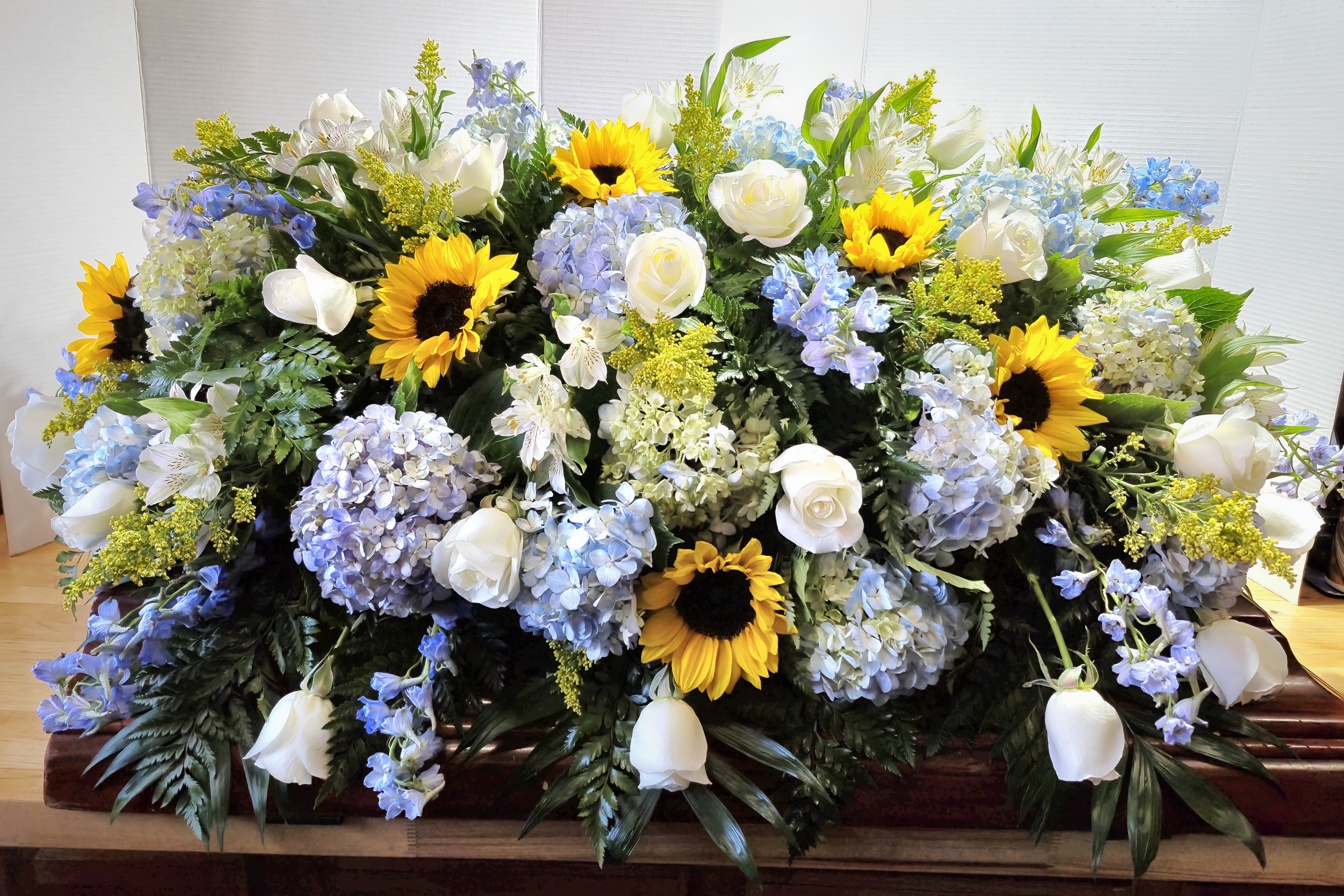 Sunflower Casket Spray  - Bright yellow sunflowers set a warm, comforting tone across this elongated casket spray, perfect for a funeral or memorial service where you want to celebrate a life with sunny, uplifting color. This sympathy arrangement combines the joyous simplicity of sunflowers with calming blue and white tones to honor the person you're remembering.  A lush, cloudlike base of blue hydrangeas and airy delphinium supports clusters of white roses and ivory alstroemeria, creating soft texture and classic form along the length of the spray. The overall composition is full and abundant, with green foliage woven throughout to balance the palette and add natural depth. The combination of flowers offers both visual warmth and serene cool accents, making the piece feel peaceful without being somber.  Made to drape gracefully on the casket, this spray is arranged for immediate presentation at services and memorials; its durable blooms are selected to maintain their appearance through viewing and ceremony. Choosing this piece sends a message of heartfelt remembrance-an arrangement that helps the recipient feel seen and comforted during a difficult time.