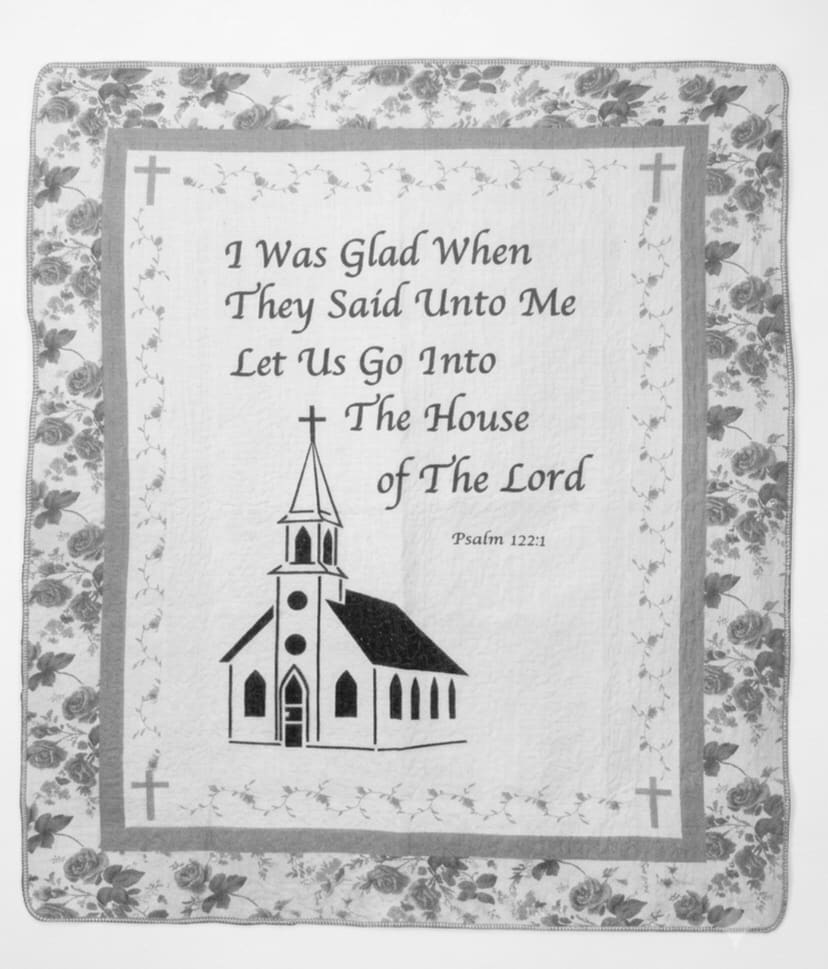 House of the Lord Psalm122:1 Throw - The devotional quilted throw brings Psalm 122:1 to life with a softly patterned rose border and a simple church illustration-perfect as a sympathy or memorial gift and for faith-based occasions like baptisms, confirmations, or remembrance services. The central panel reads "I Was Glad When They Said Unto Me Let Us Go Into The House of The Lord" above the church image, so the person you're honoring will feel remembered and comforted by a tangible expression of faith.  This 50" x 60" fabric wall-hanging/throw is lightweight, machine washable, and built for everyday use or display. Muted whites, light grey and grey accents give it a traditional look; the quilted texture and printed text create an heirloom quality that suits a mantel, pew, or chair. A matching fabric bow is included for presentation, and the piece pairs well with the optional wire easel stand for upright display at a memorial or inside a church setting.